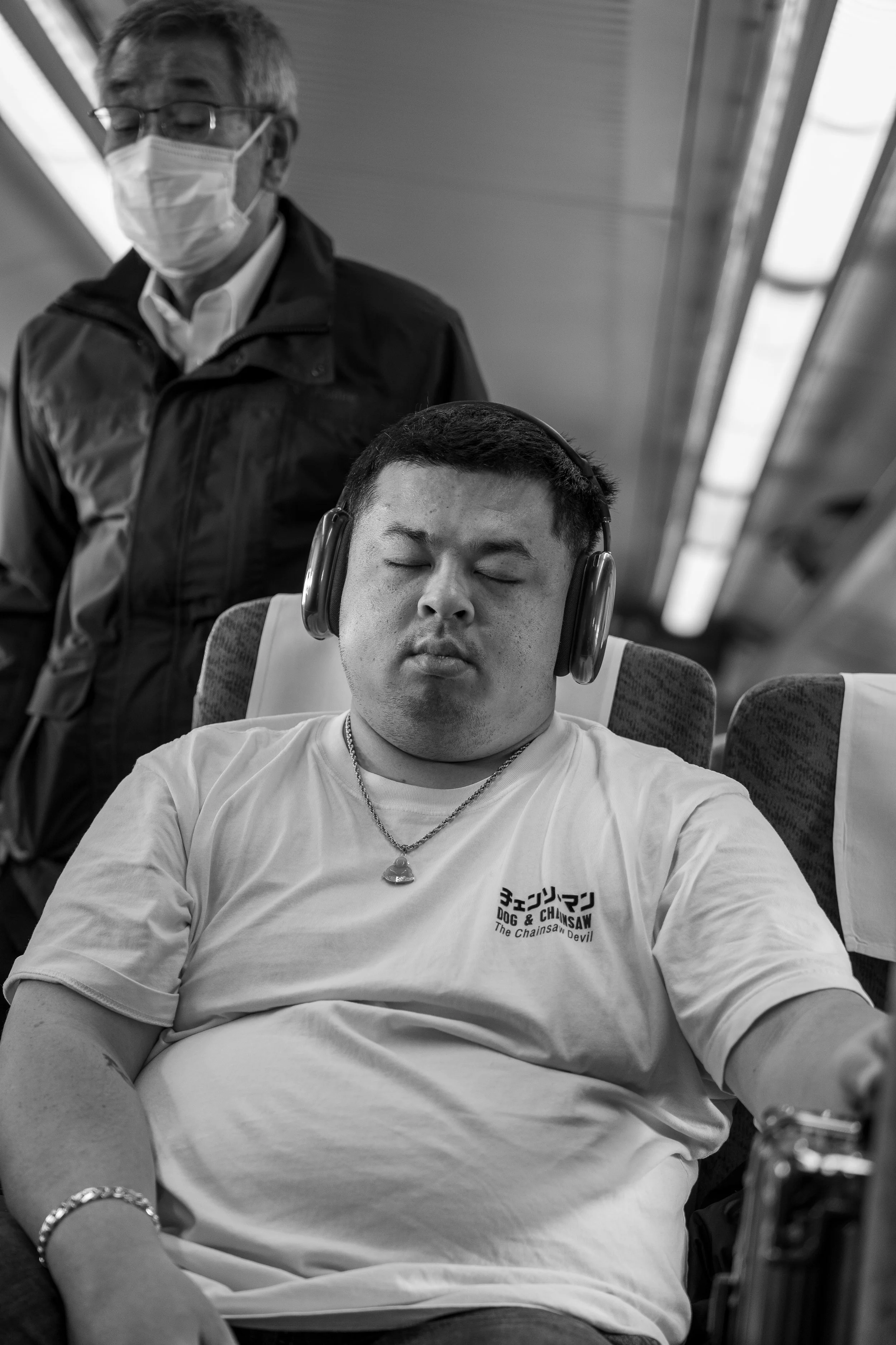 A young man with headphones sleeping on an airplane, wearing a white t-shirt with Japanese characters and English text, with an older man standing nearby wearing glasses, a face mask, and a jacket.