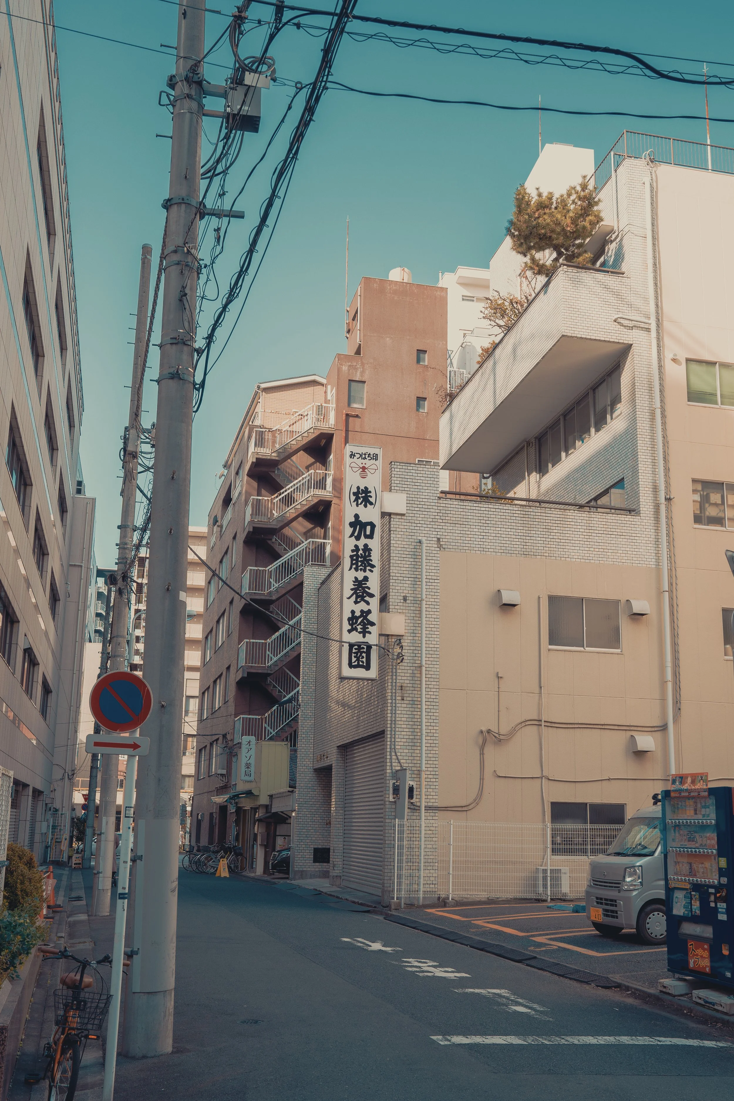 A narrow urban street with buildings, a utility pole with wires, a no parking sign, and a small truck parked on the side, under a clear blue sky.