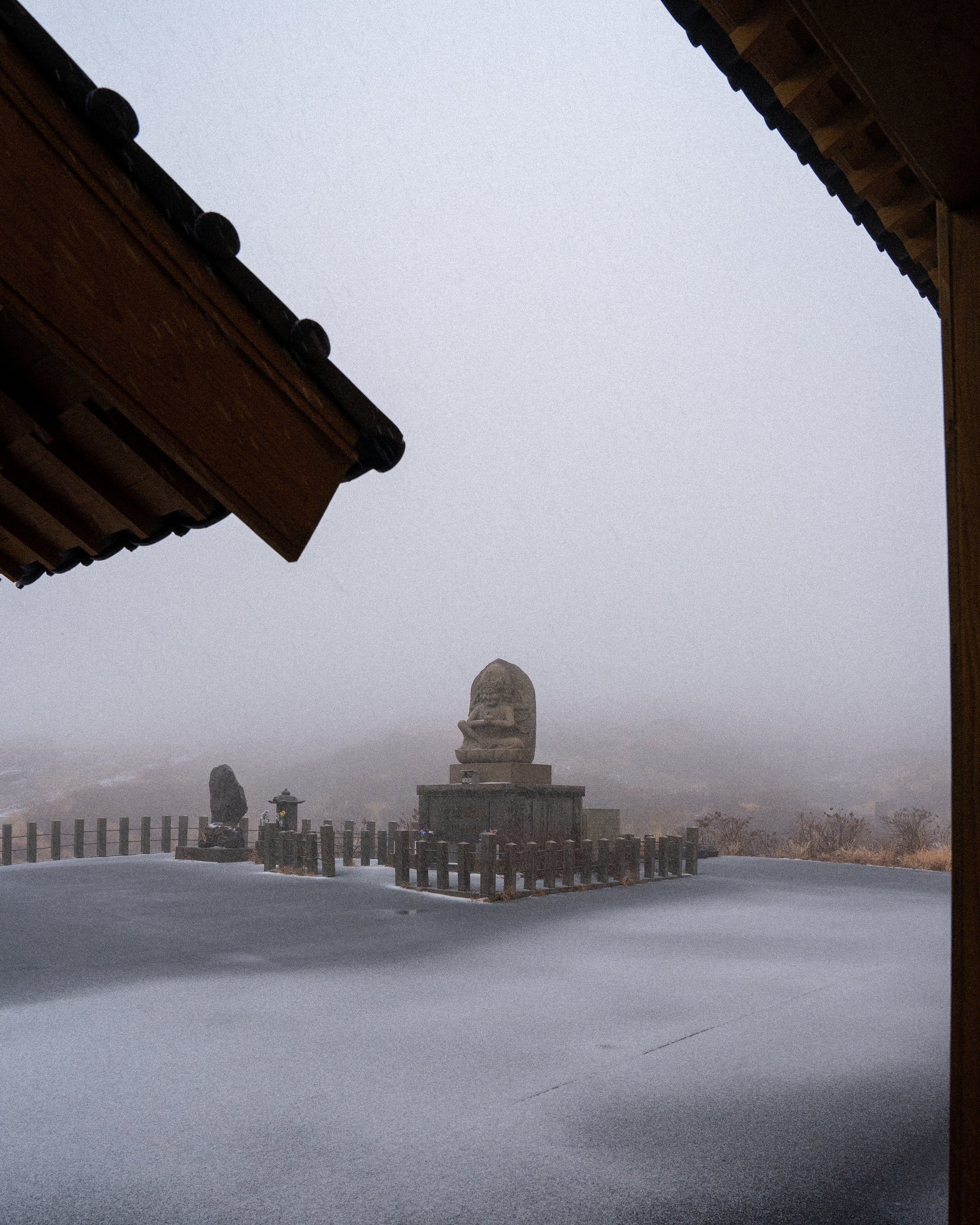 Snow-covered courtyard with Buddhist statue and lanterns, framed by traditional wooden roof edges, foggy background