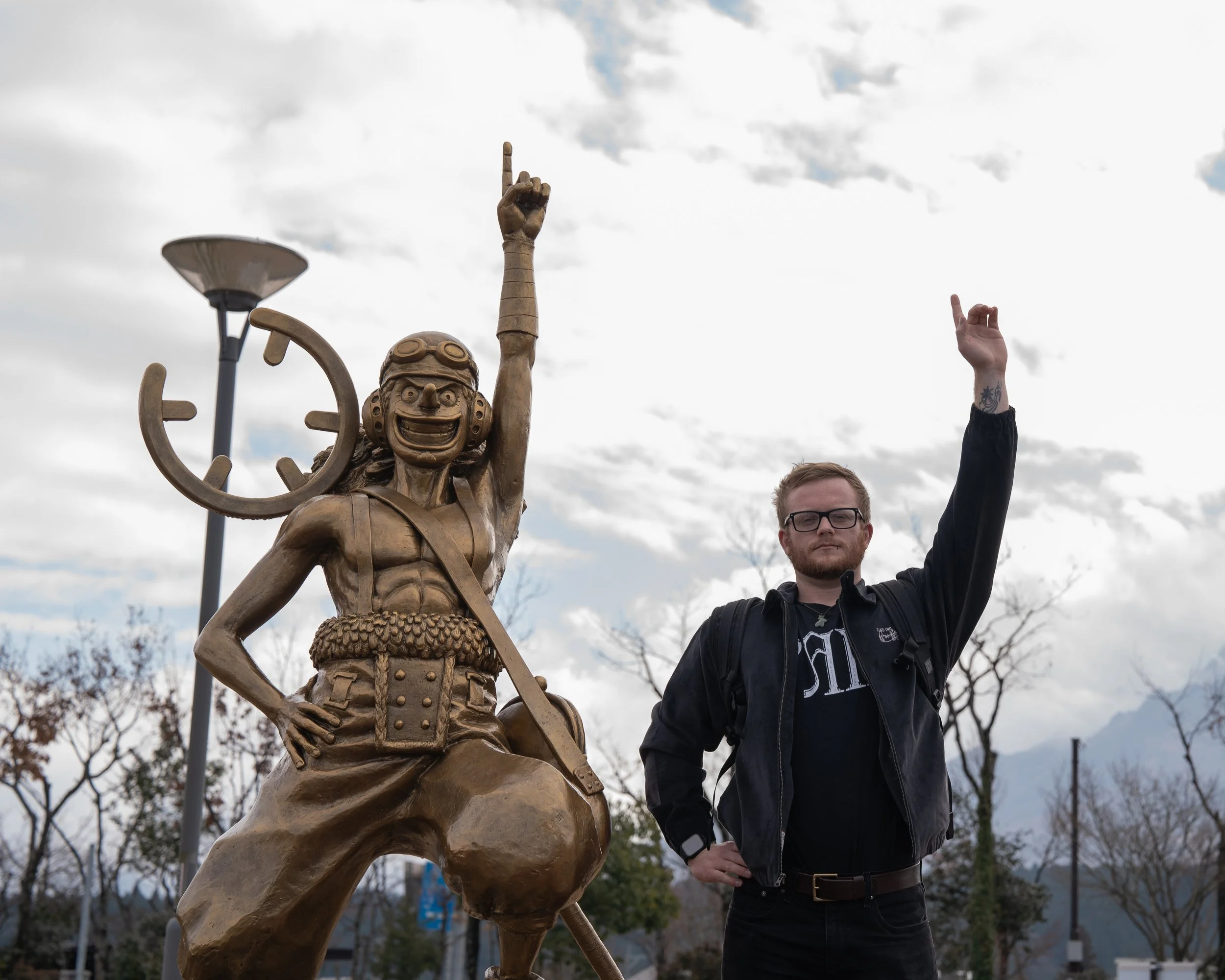 A man with glasses and a beard standing next to a large bronze statue of a cartoonish character with a big smile, goggles, and a clock on its back, both raising one hand in a fist. The sky is cloudy and there are trees and mountain peaks in the backg