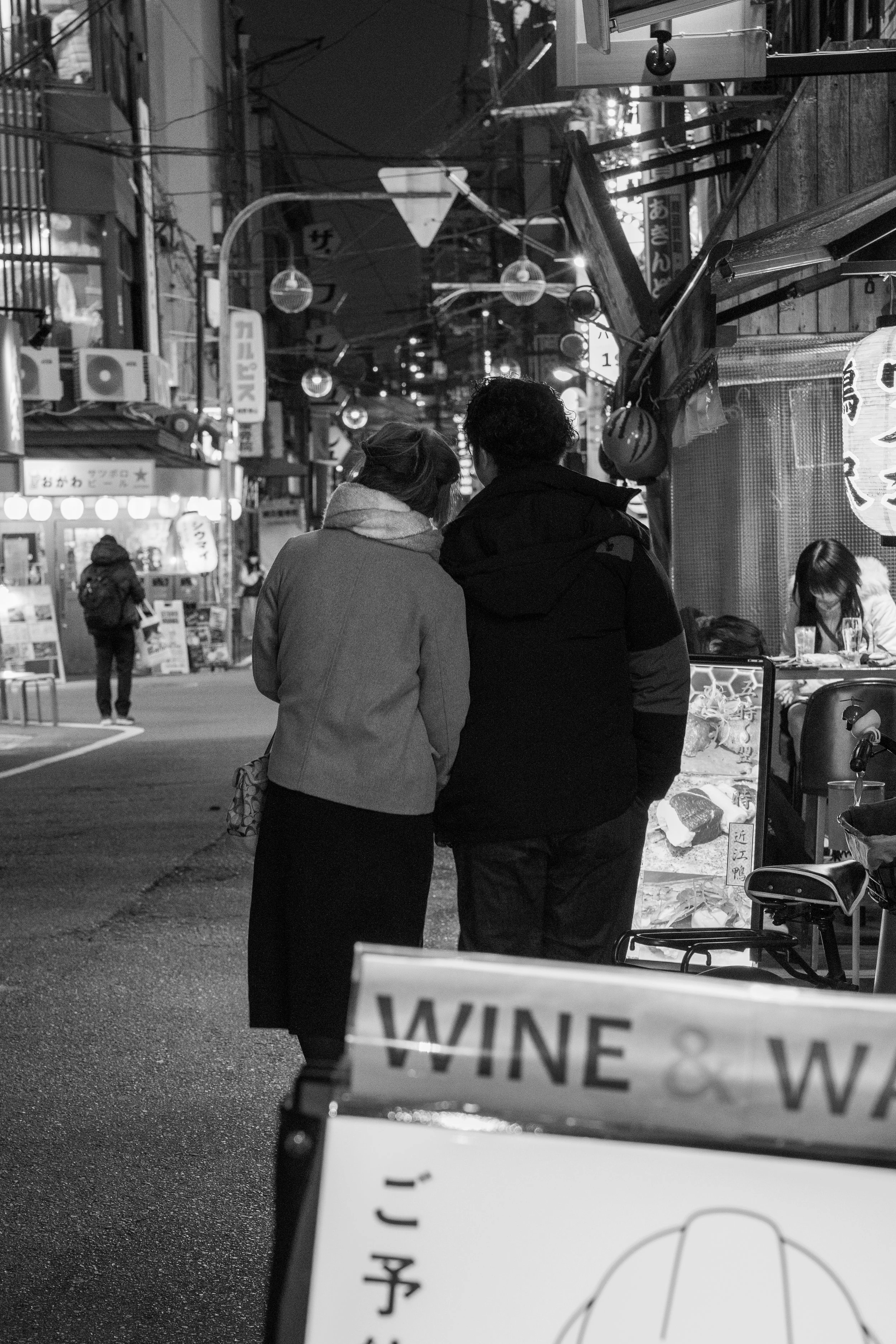 Two people stand close together on a city street at night, viewing a shop menu that is illuminated. The scene is bustling with neon signs and hanging lanterns, indicating a lively urban nightlife environment.