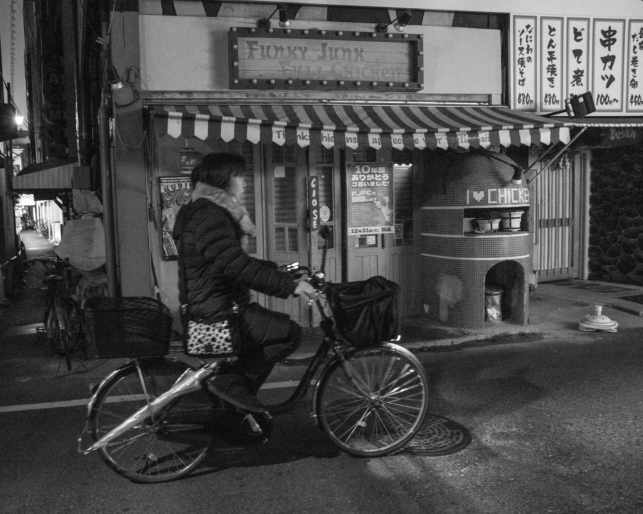 A person riding a bicycle on a city street at night, with a restaurant or cafe in the background. The scene is in black and white.