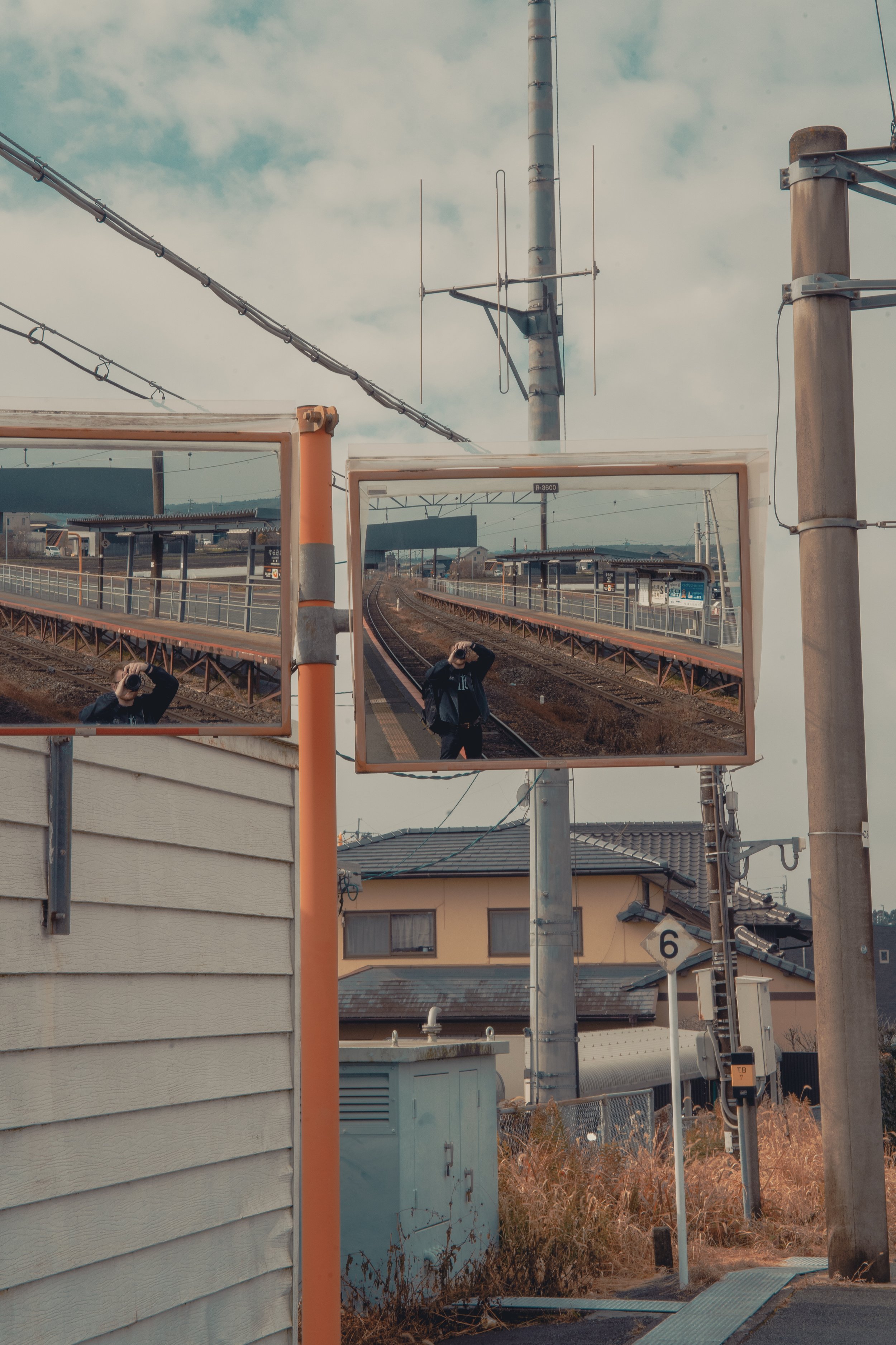 A person taking a photograph with a camera, seen through two mirrors on a street near a train station, with railway tracks, a blue sky with clouds, and houses in the background.