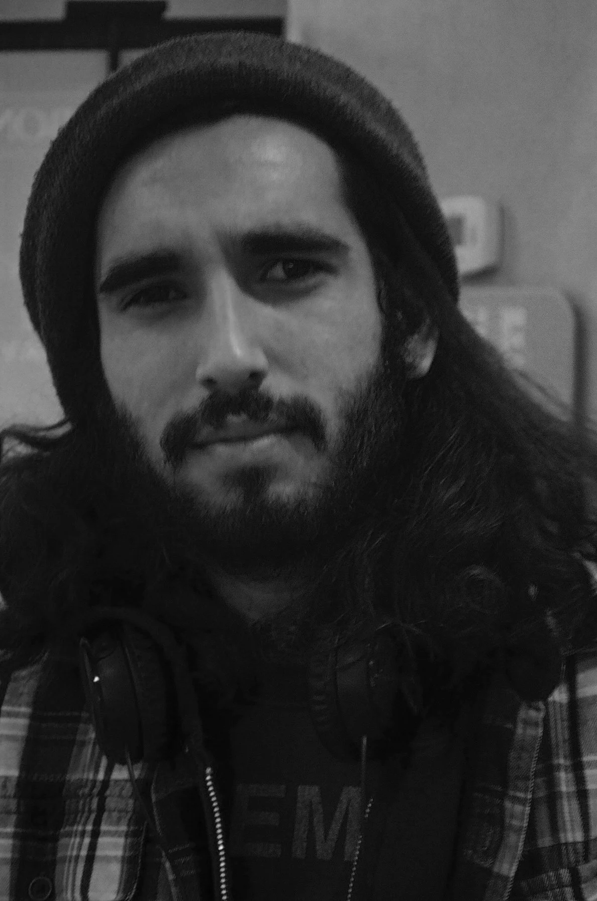 Black and white photo of a young man with long hair, beard, and mustache, wearing a beanie, headphones around his neck, and a plaid shirt over a t-shirt.