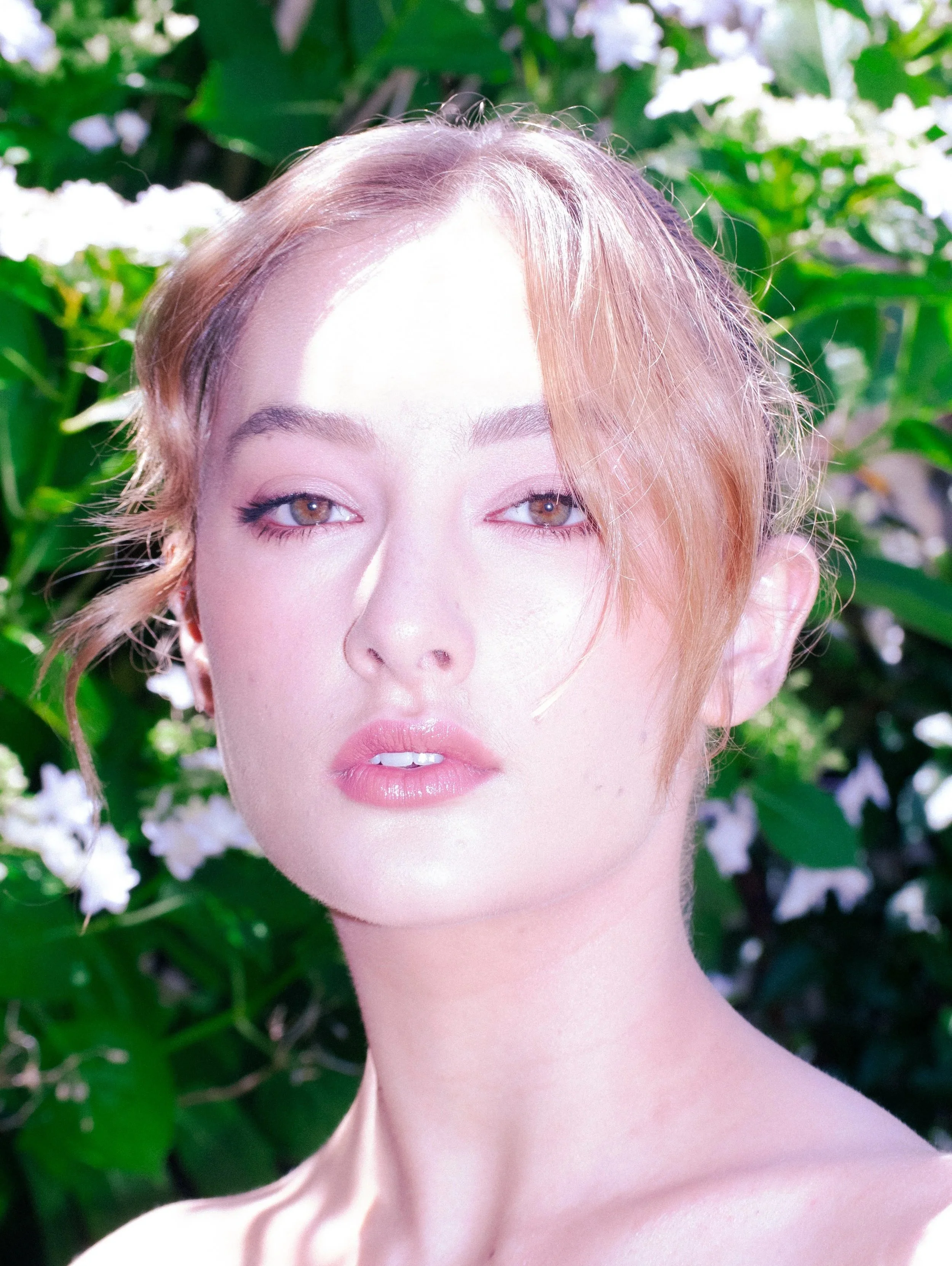 Young woman with romantic soft glam makeup, subtle smokey eyeliner, outdoors with greenery and white flowers.