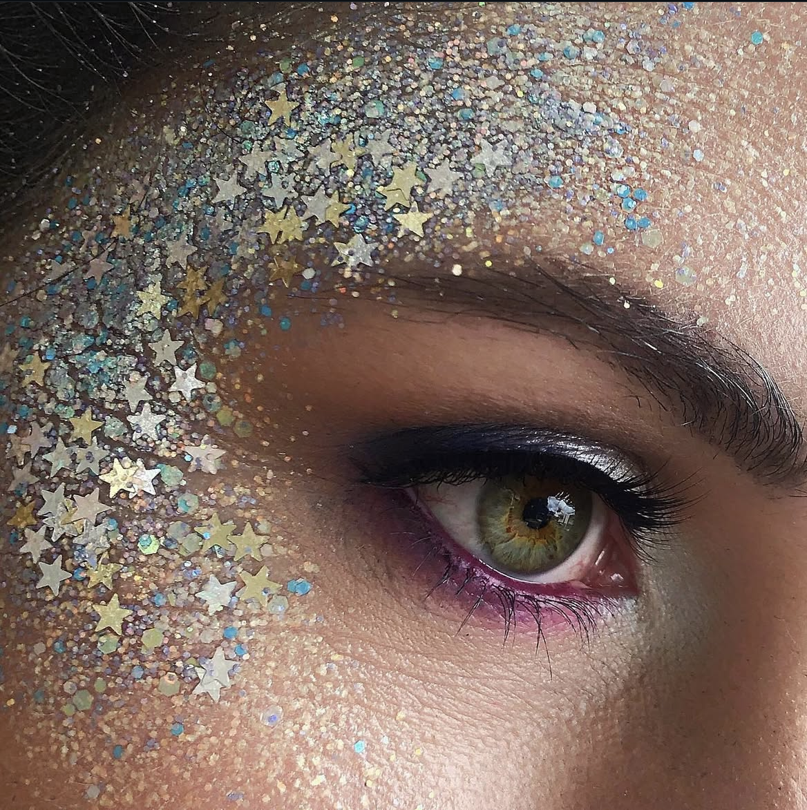Close-up of eye with colorful glitter and star confetti, featuring dark eyeliner and shimmery eyeshadow for a festival or creative makeup look.