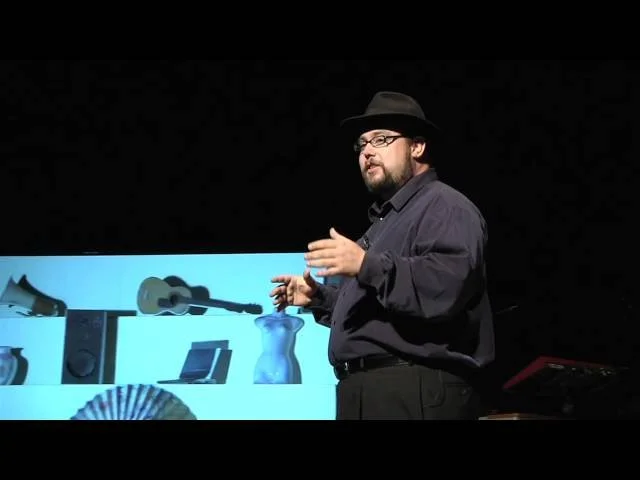 Man wearing a hat and glasses giving a presentation with various objects displayed on a screen in the background.