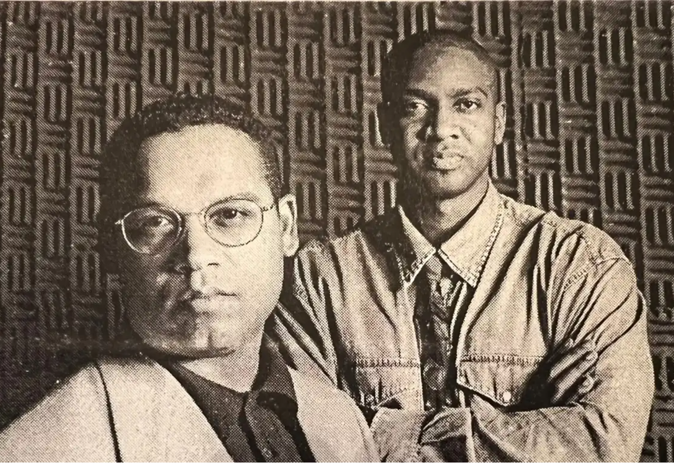 Two men in front of a patterned wall, one wearing glasses and a suit, the other in a denim shirt.
