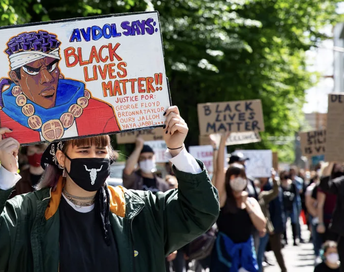 Protesters holding Black Lives Matter signs, including one with an anime drawing, at a demonstration. Trees are visible in the background.