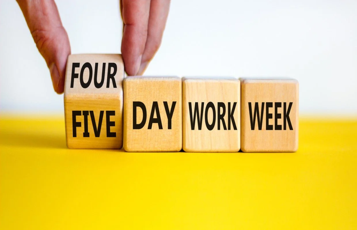 Wooden blocks on a yellow background displaying 'Four Day Work Week' with a hand adjusting the blocks.