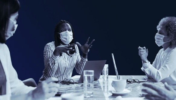Group of diverse people wearing masks engaged in a discussion around a table with laptops and coffee cups in a professional setting.