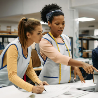 Two women wearing work aprons reviewing documents and using a laptop in a workshop setting.
