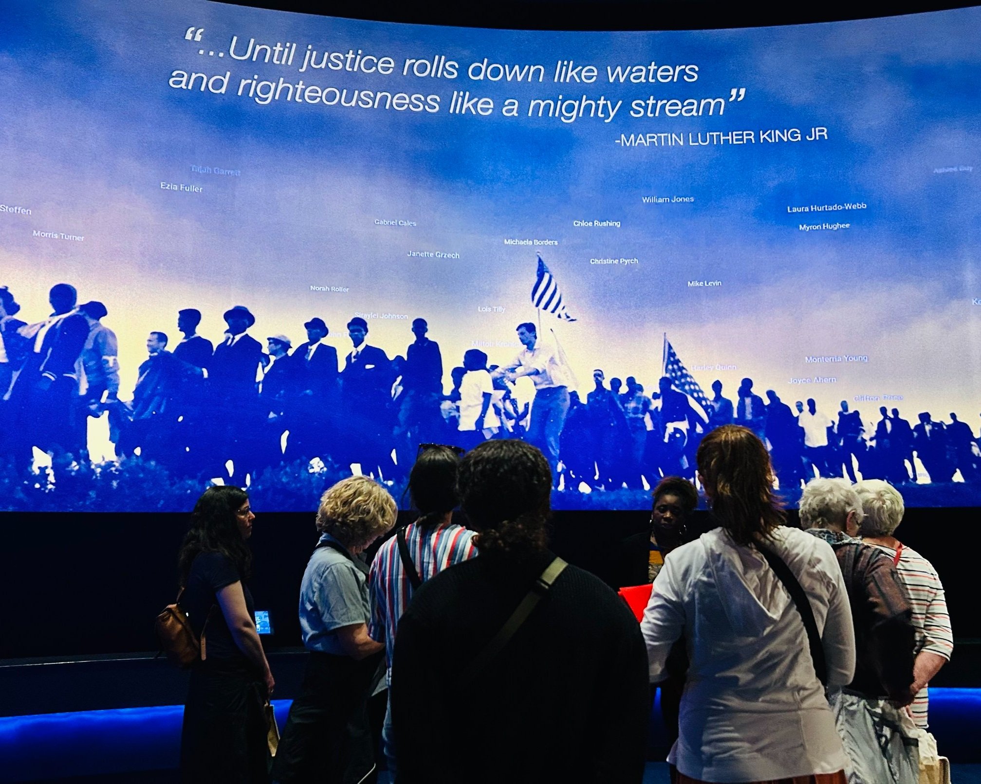 People viewing a large screen displaying a quote by Martin Luther King Jr., an image of a march with people holding American flags, and various names over the sky background.