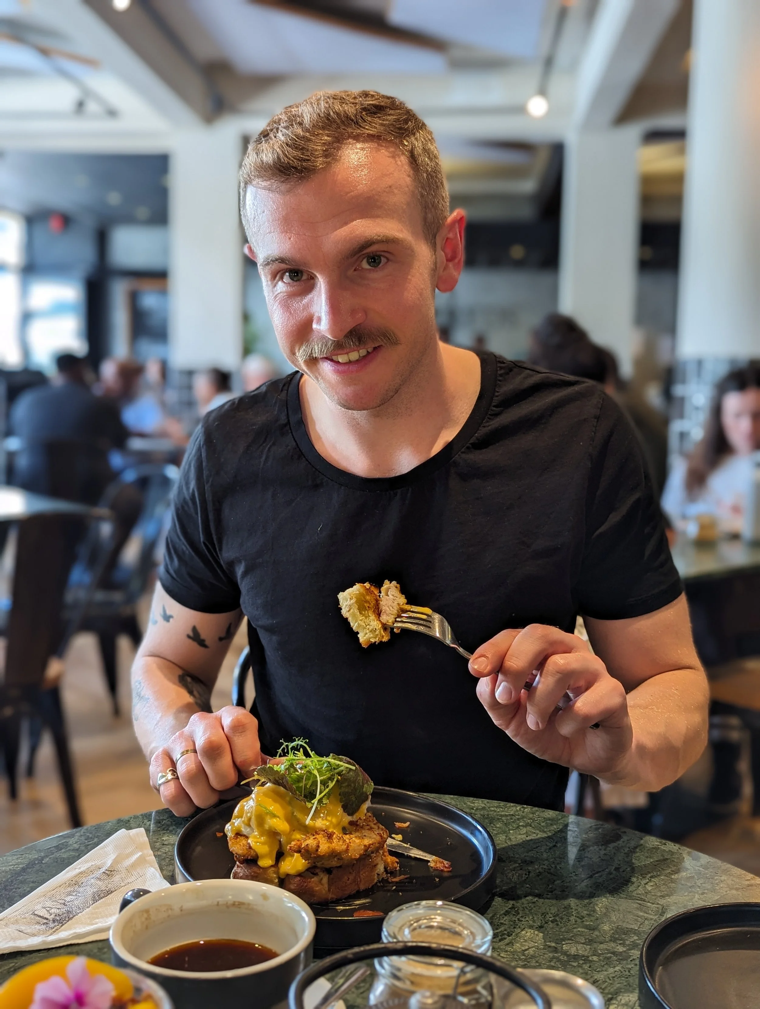 Person in a black shirt seated at a restaurant table with a plate of food, eating with a fork. The dish includes bread, egg, and greens. Coffee cup and napkin on the table. Background shows other diners.