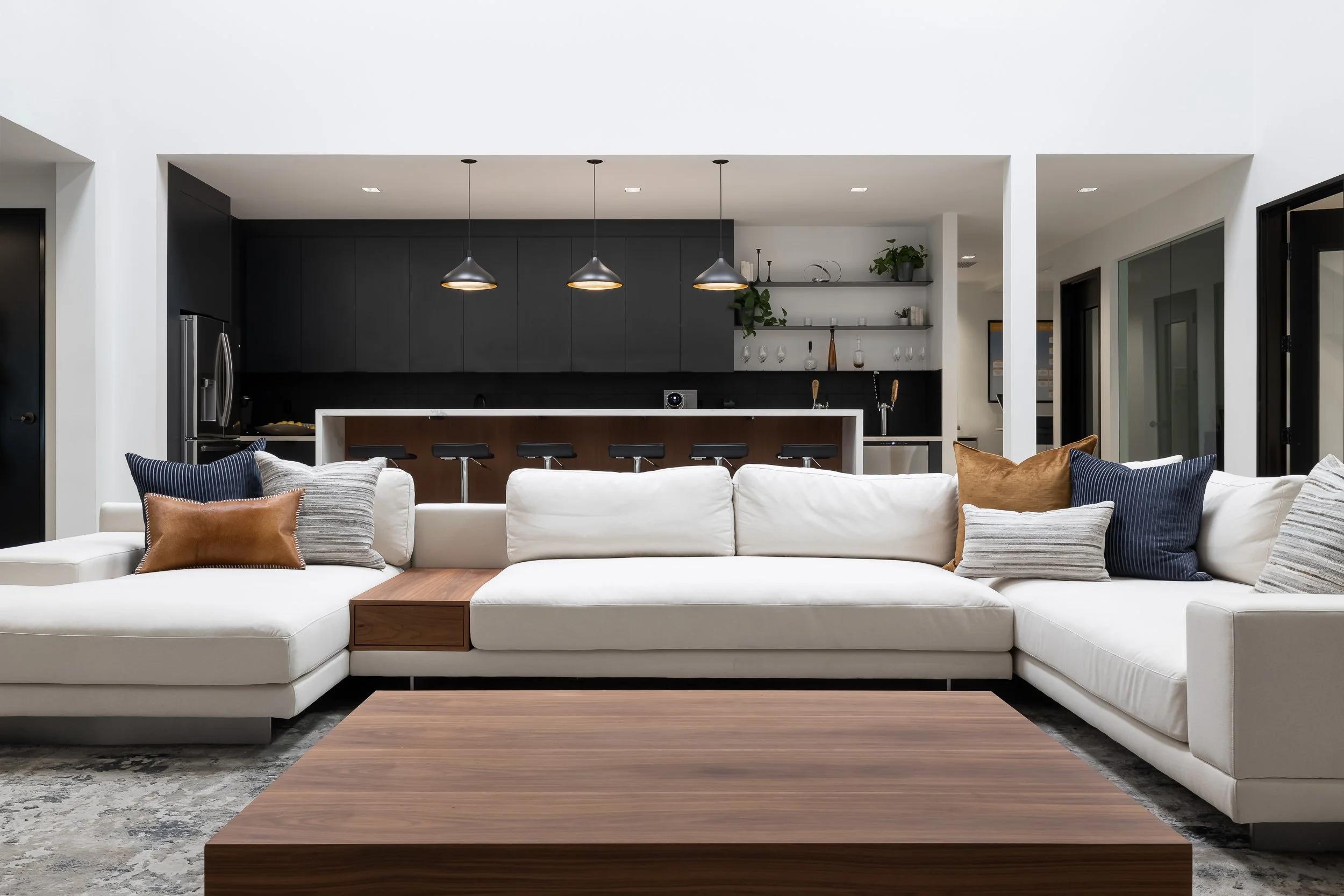 A modern open-plan living room with a white sectional sofa, decorative pillows, and a wooden coffee table. The background features a black kitchen island with barstools, black cabinets, and a black wall with open shelves and plants. Modern pendant li