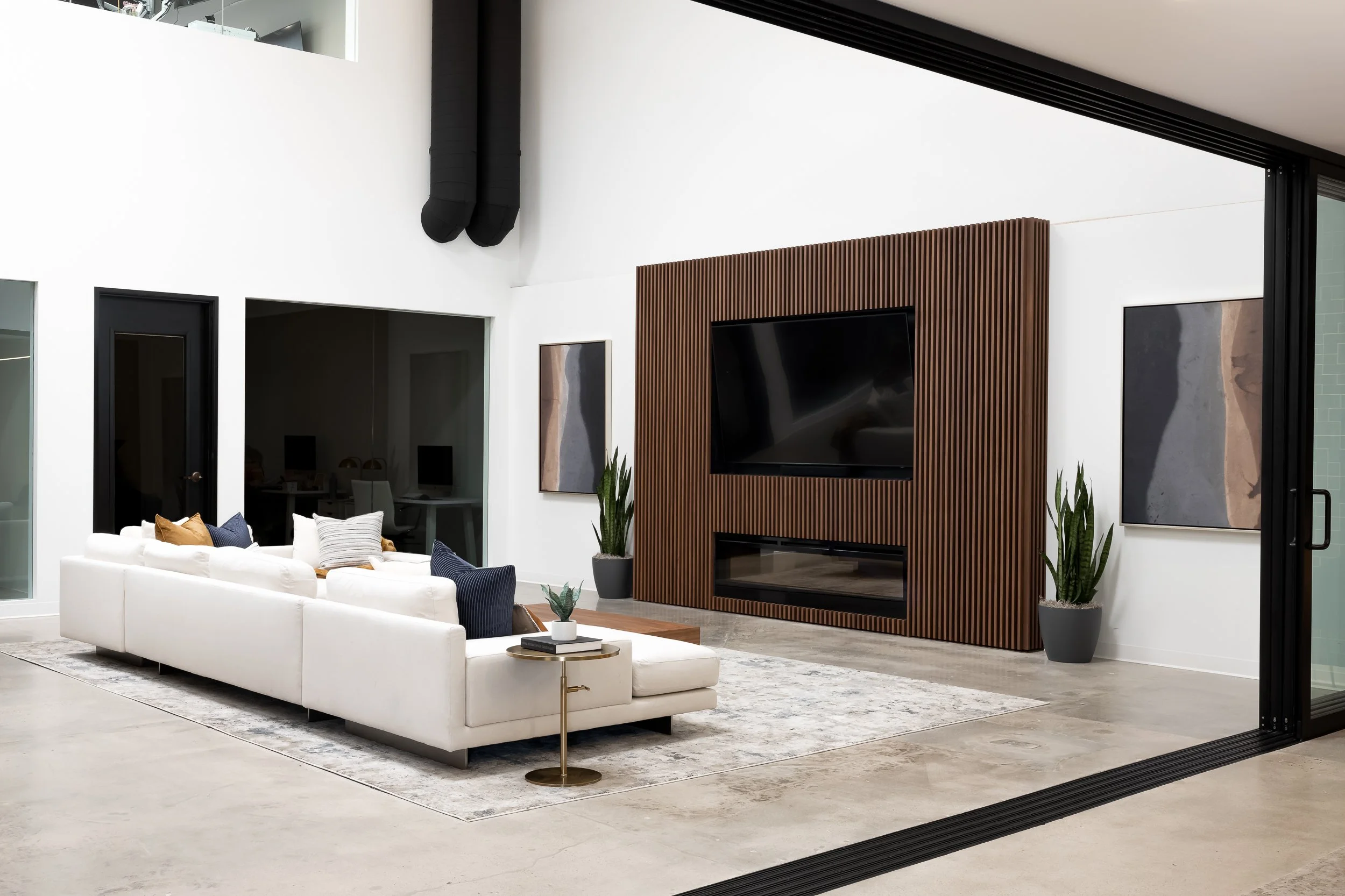 Modern living room with white sectional sofa, decorative pillows, and a small side table. A large flat-screen TV is mounted on a wooden slatted wall, flanked by tall plants. The room features high ceilings, black pipes, and glass sliding doors.