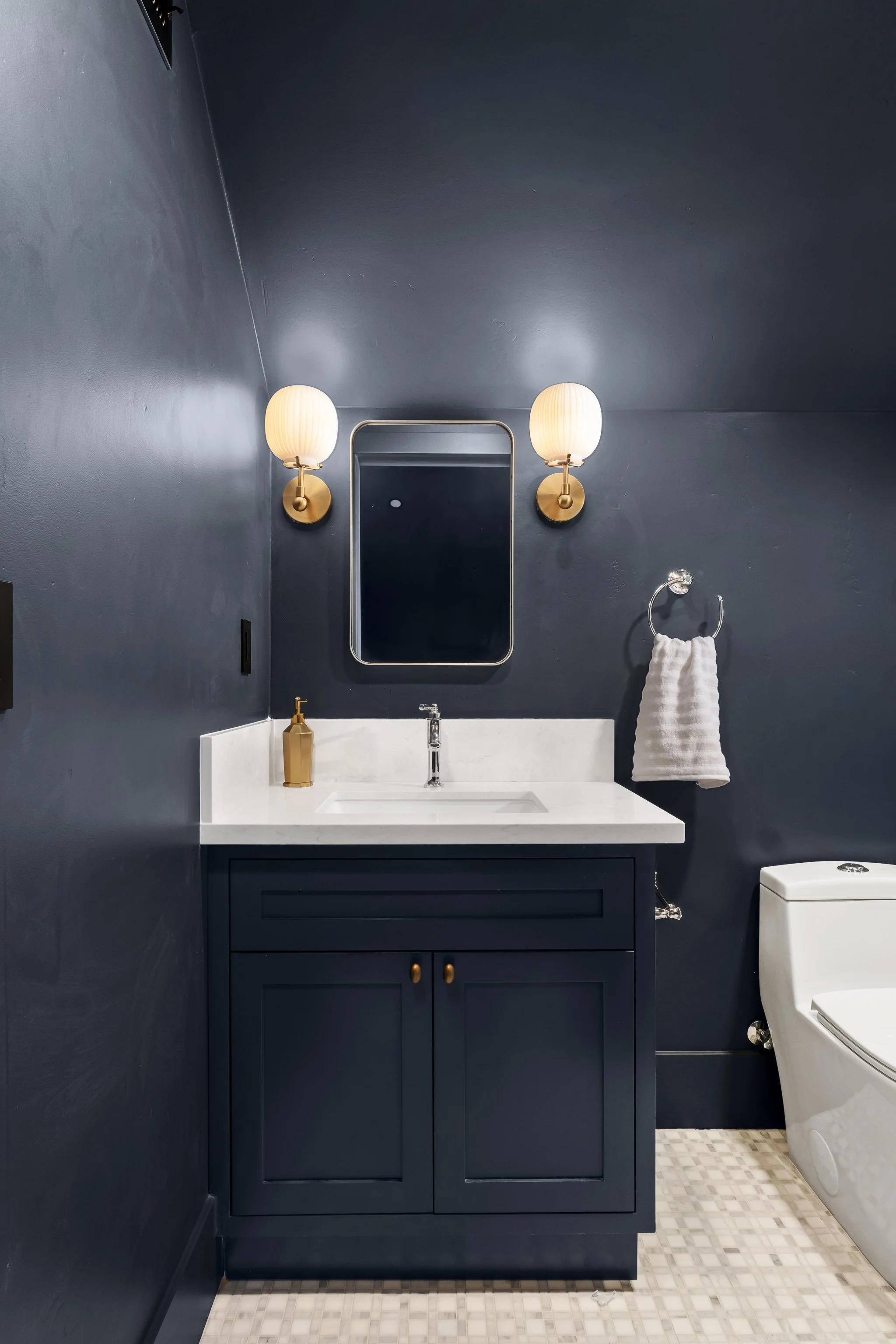 A modern bathroom with dark blue walls, a white countertop with a sink, a gold soap dispenser, and two wall-mounted lamps with shaded lights. There is a rectangular mirror above the sink, a towel hanging on a ring, and a white toilet to the right.