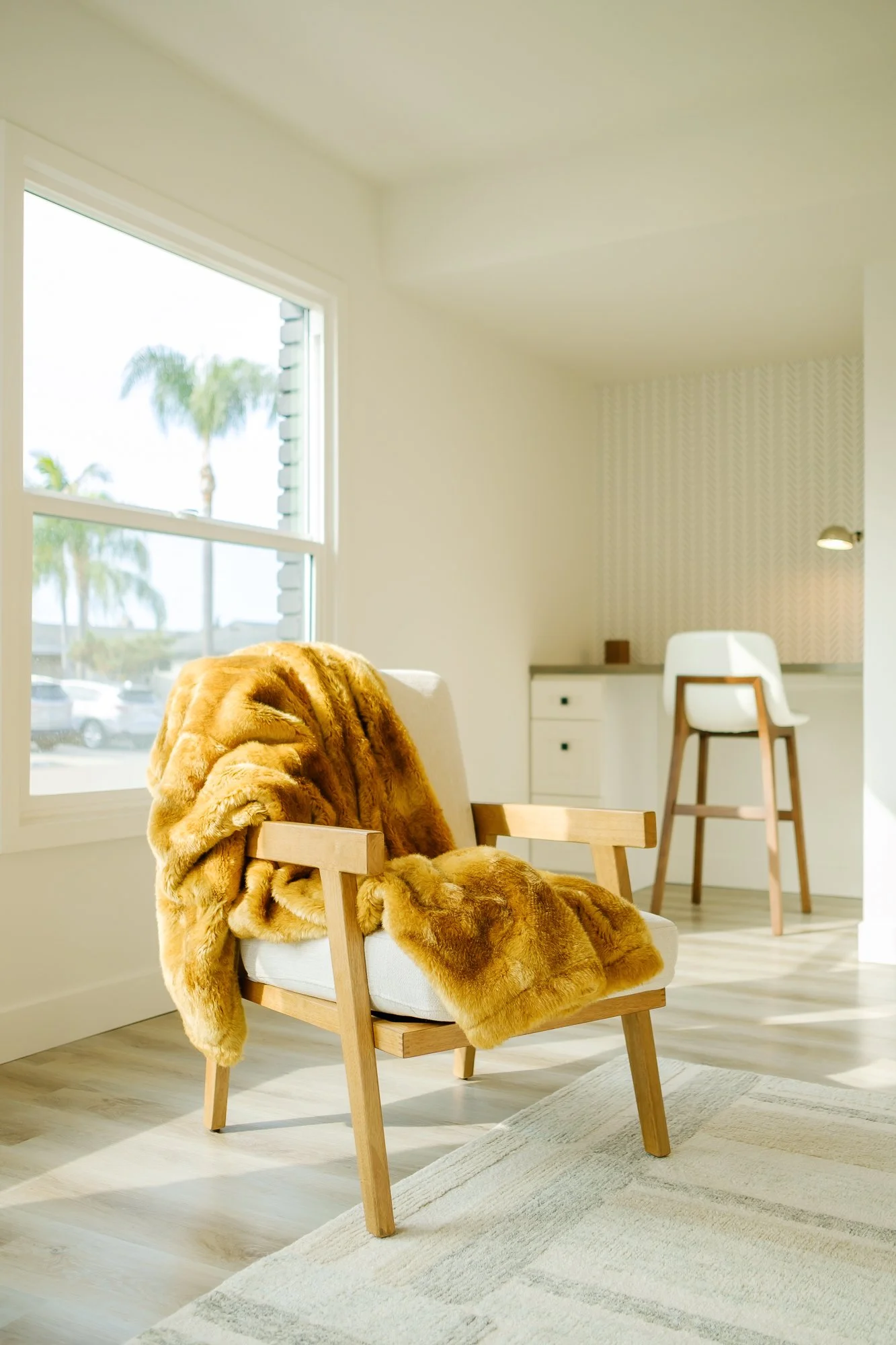 Bright living room with a wooden chair draped with a large, plush, golden-brown faux fur blanket, near a window with a view of palm trees and a parking lot, with a small desk and white chair in the background.