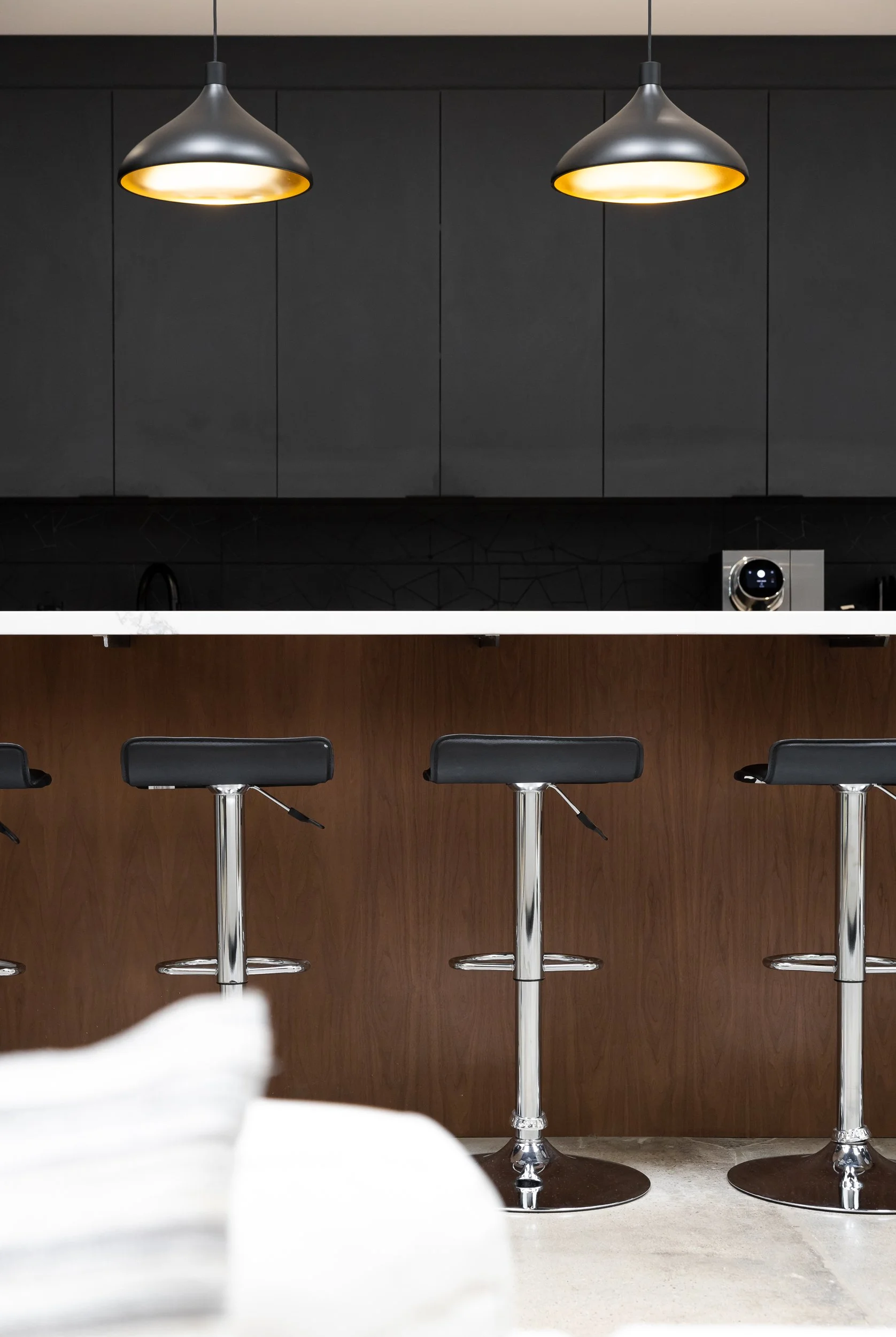Interior of a modern space with two black pendant lights hanging from the ceiling, black bar stools with chrome bases aligned in front of a bar or counter with a wood panel and black wall behind it, and a coffee machine on the right side.
