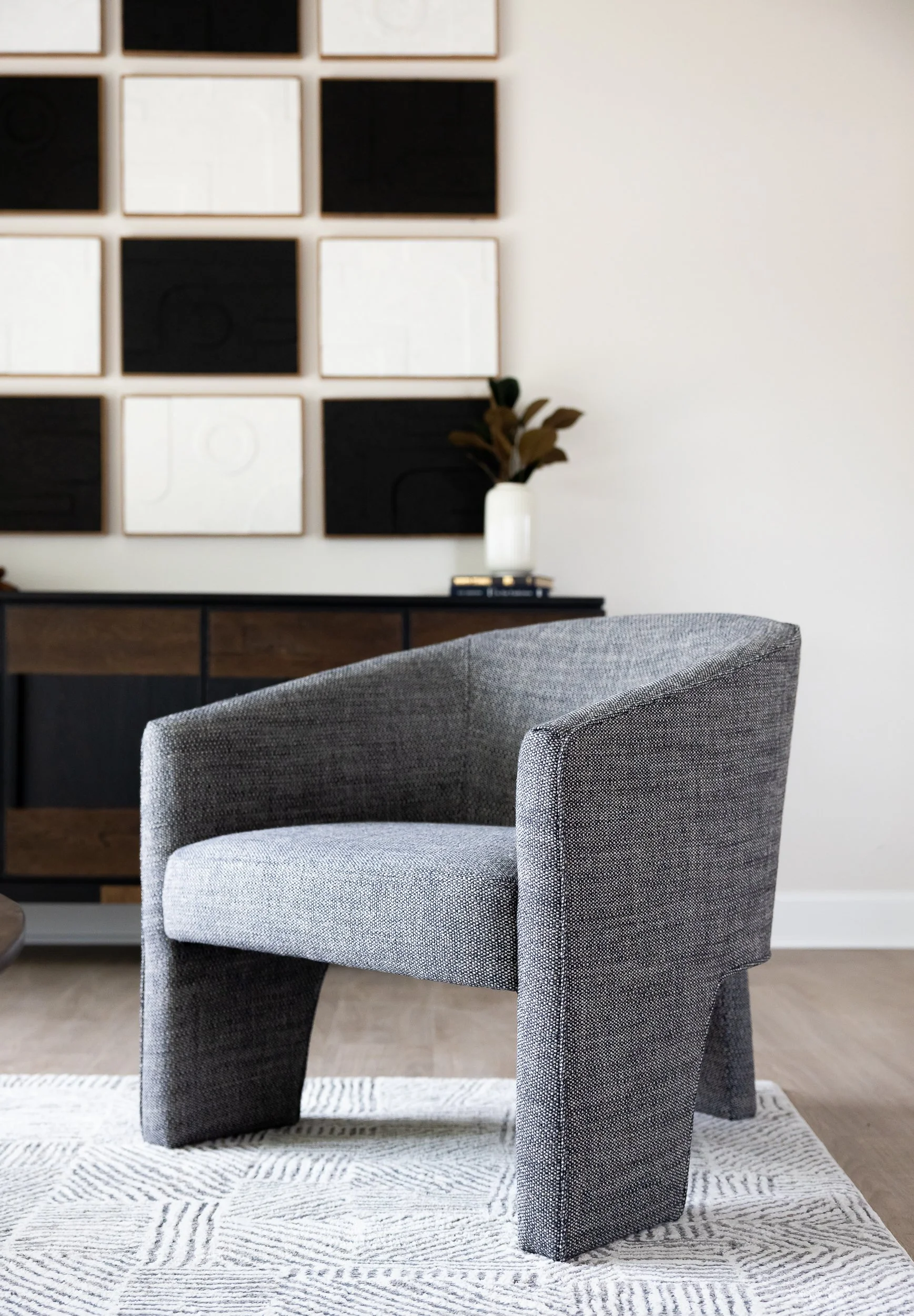 Gray upholstered armchair with curved armrests on a light patterned rug in a modern living room. In the background, a wooden sideboard with a white vase holding brown leaves and a stack of books, and a black and white grid artwork on the wall.