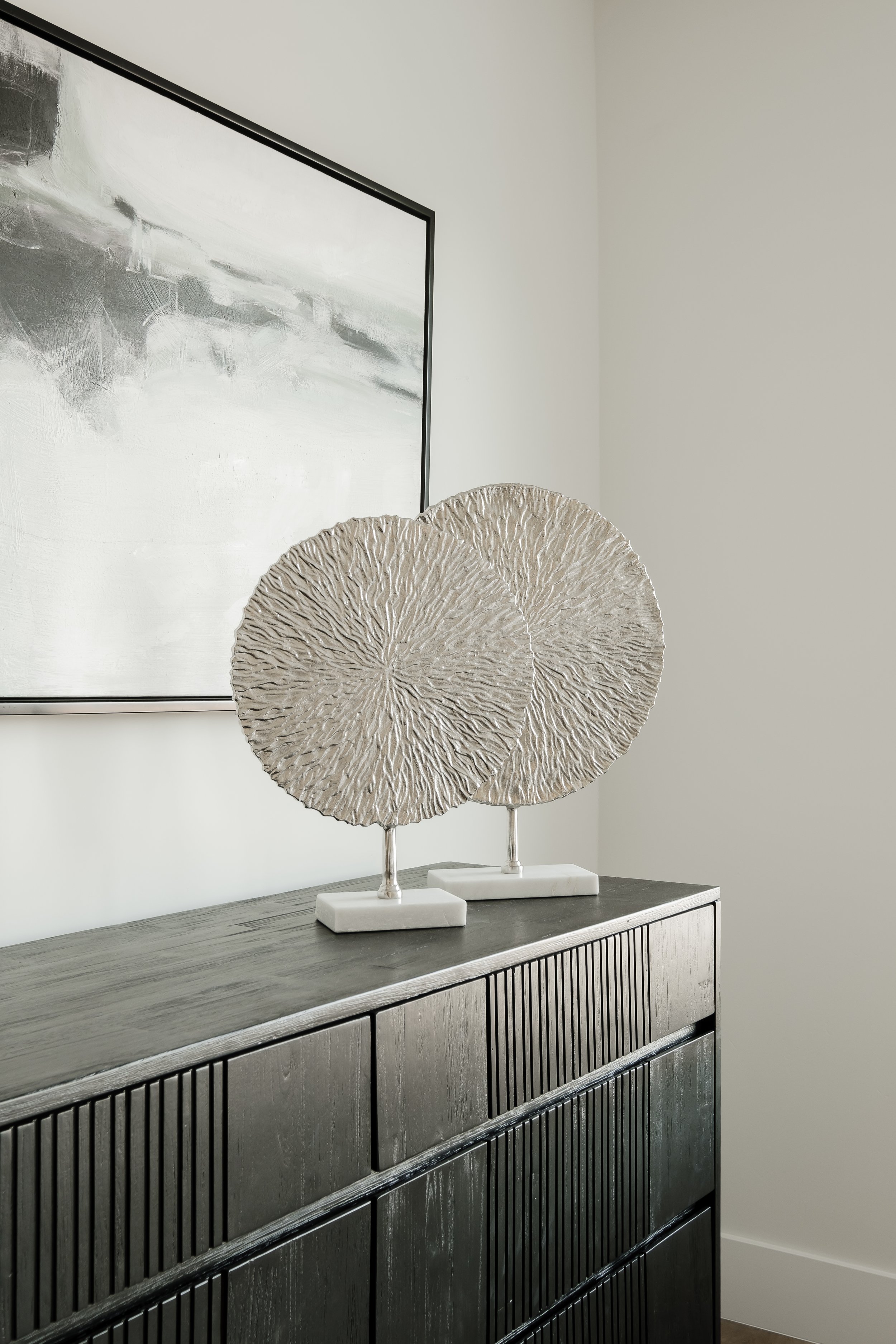 Decorative metallic sculptures resembling large, textured, circular leaves placed on white stands on a black wooden sideboard against a light-colored wall with abstract artwork.