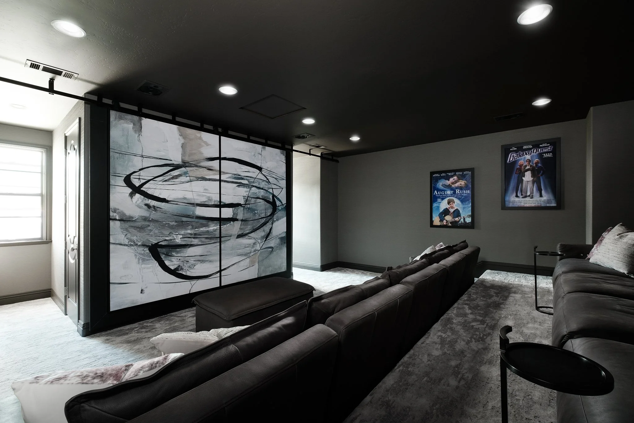 A living room with a black ceiling ceiling, recessed lighting, and framed movie posters on the wall. There is a black abstract artwork on a divider, sitting in front of a window with blinds. The room has gray carpeting, a gray sofa, and pillows.