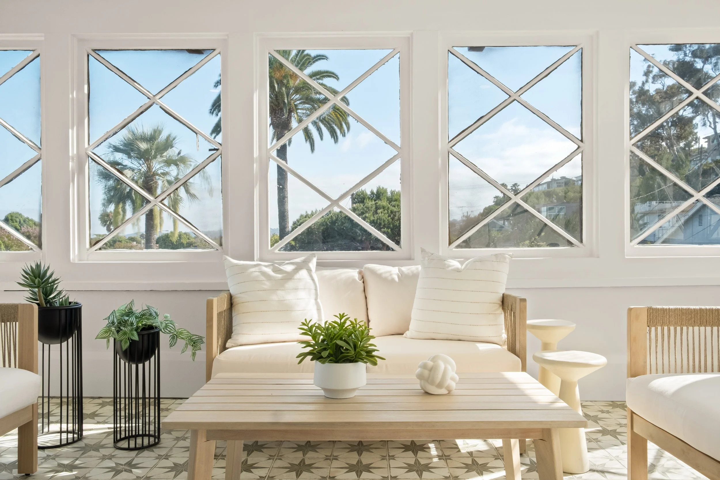 Bright living room with large windows showing palm trees and blue sky outside. White sofa with striped cushions, wooden coffee table with white decorative objects, and plants in black pots. Tiled floor with geometric pattern.