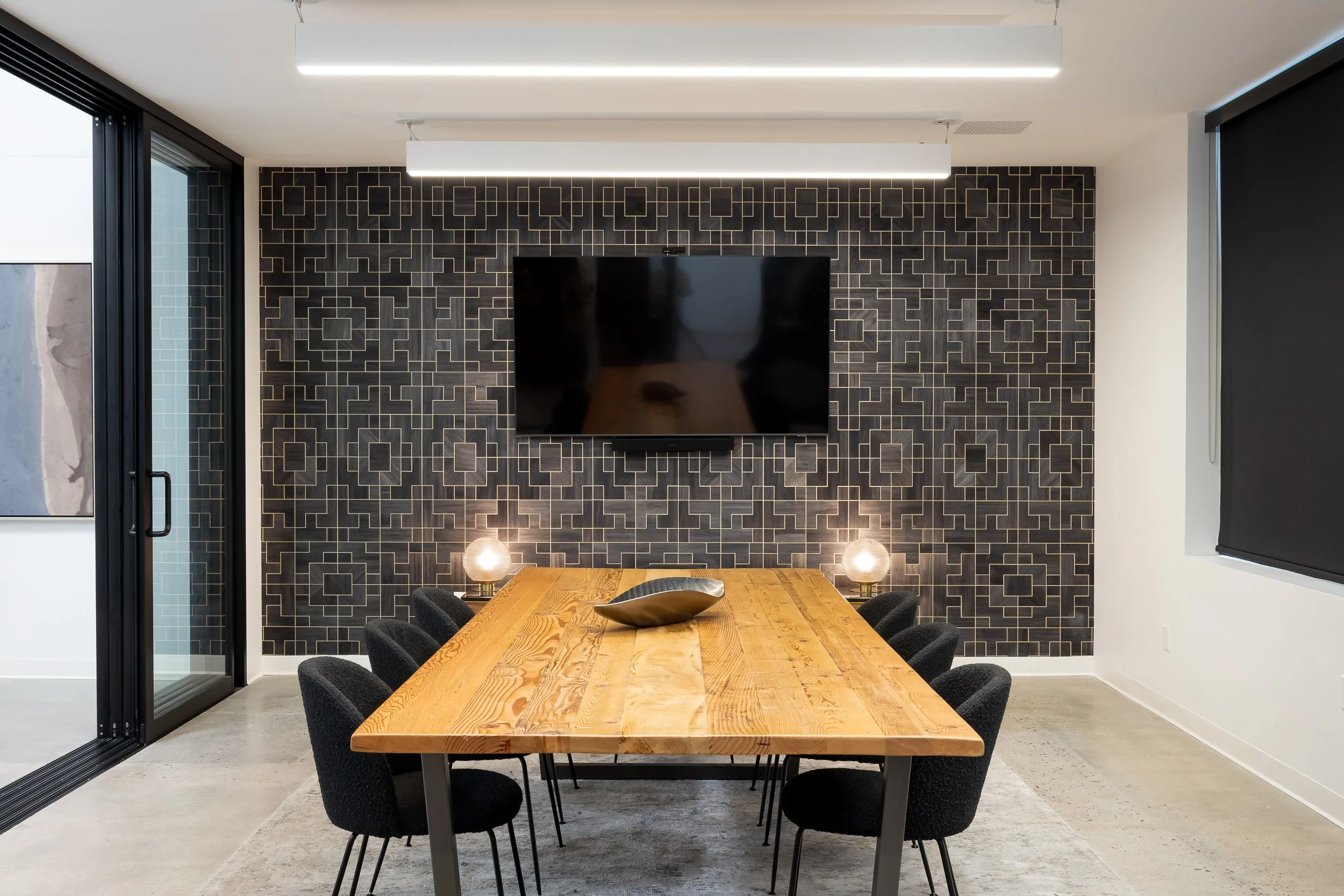 Modern conference room with wooden table, black chairs, black patterned accent wall, and two small table lamps, with sliding glass door on the left and window with black roller shade on the right.