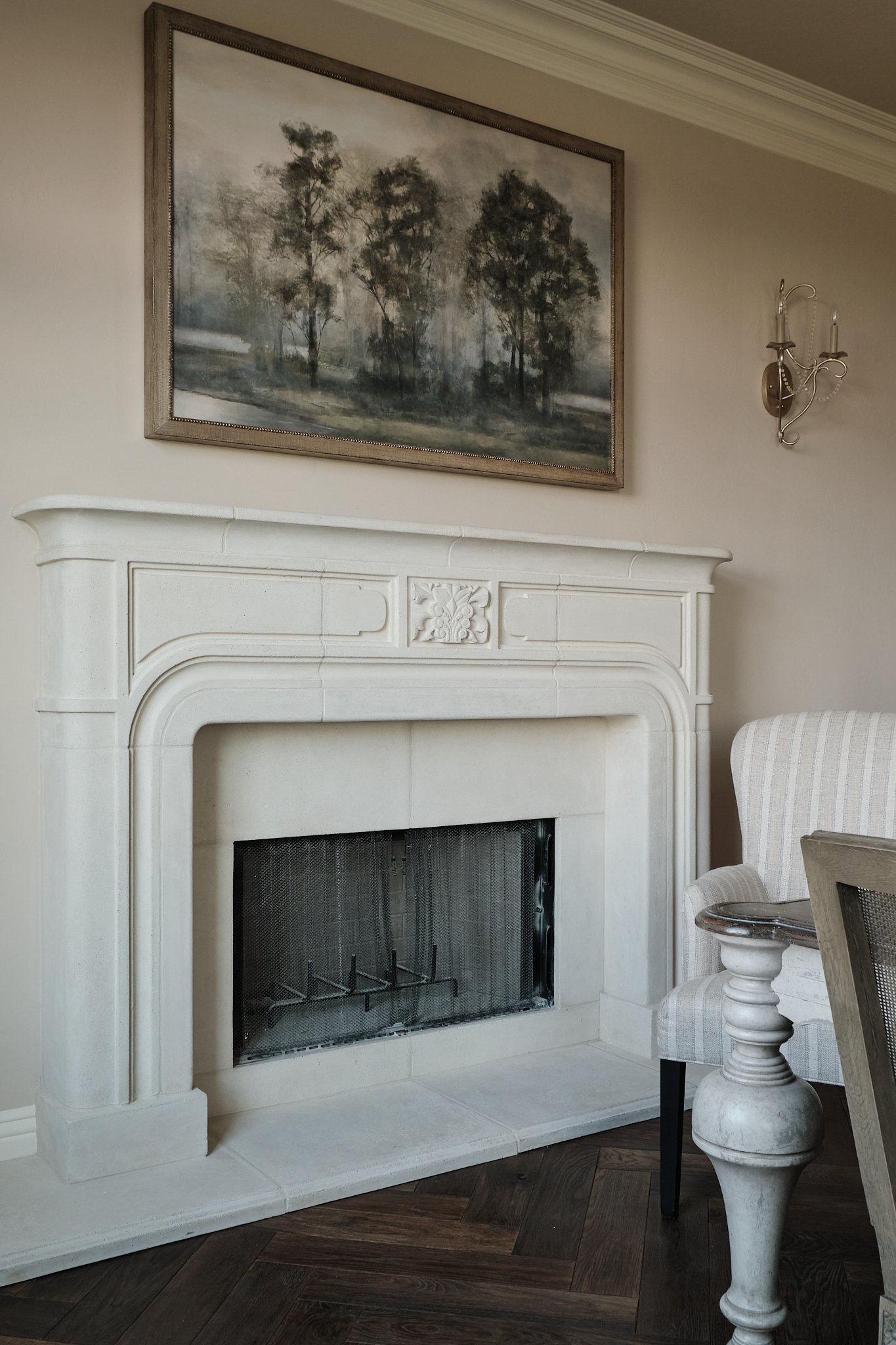A white fireplace with a decorative mantel and a wire fire screen, next to a beige and white striped armchair and a small round marble-top side table with a classical-style base, in a room with hardwood flooring and a wall-mounted sconce light fixtur