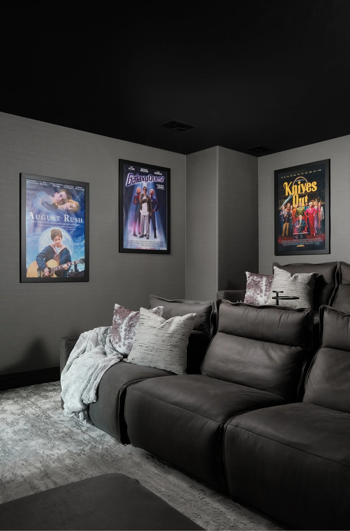 A cozy living room corner with a gray sectional sofa adorned with decorative pillows and a throw blanket, set against a dark gray wall decorated with movie posters.