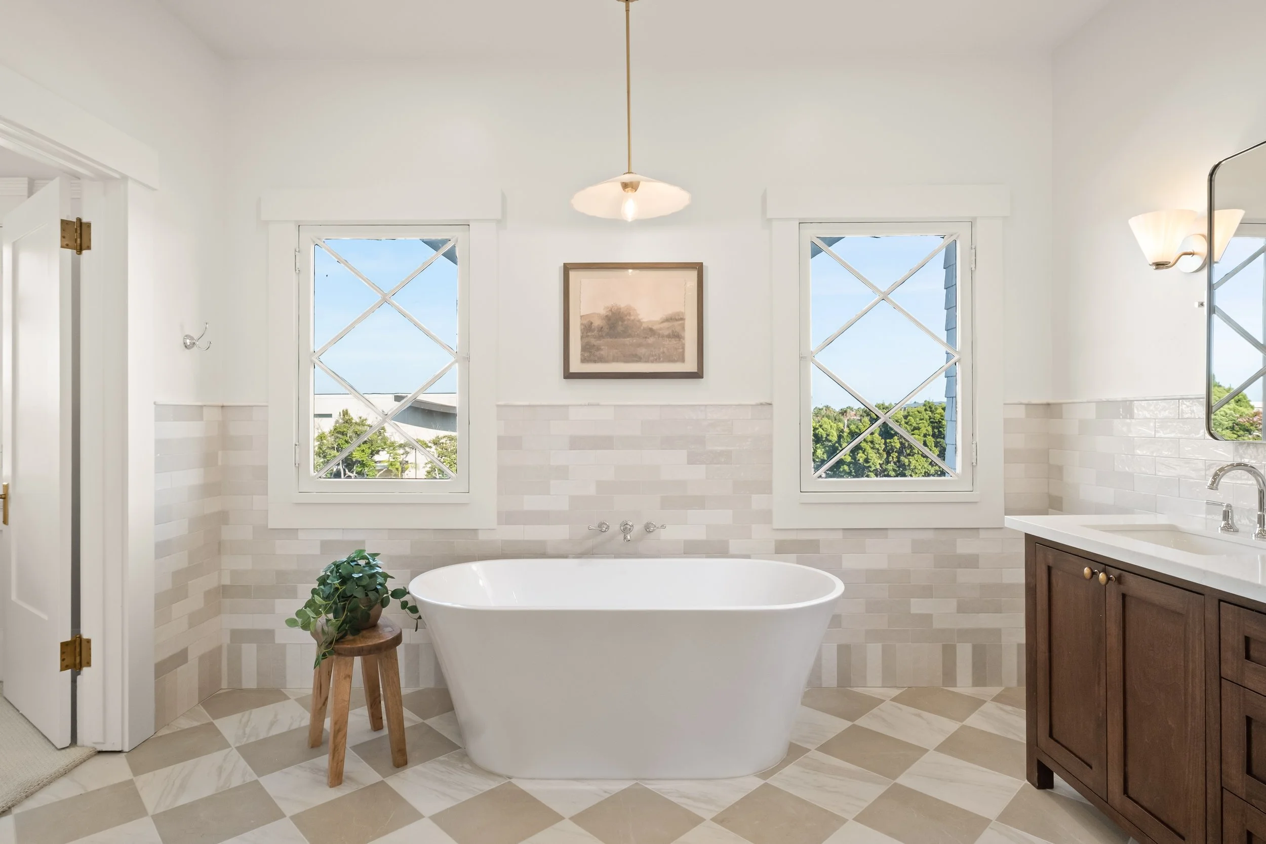 Bright bathroom with two large windows, a freestanding bathtub, a wooden stool with a potted plant, and a vanity with a dark wood cabinet and white countertop.