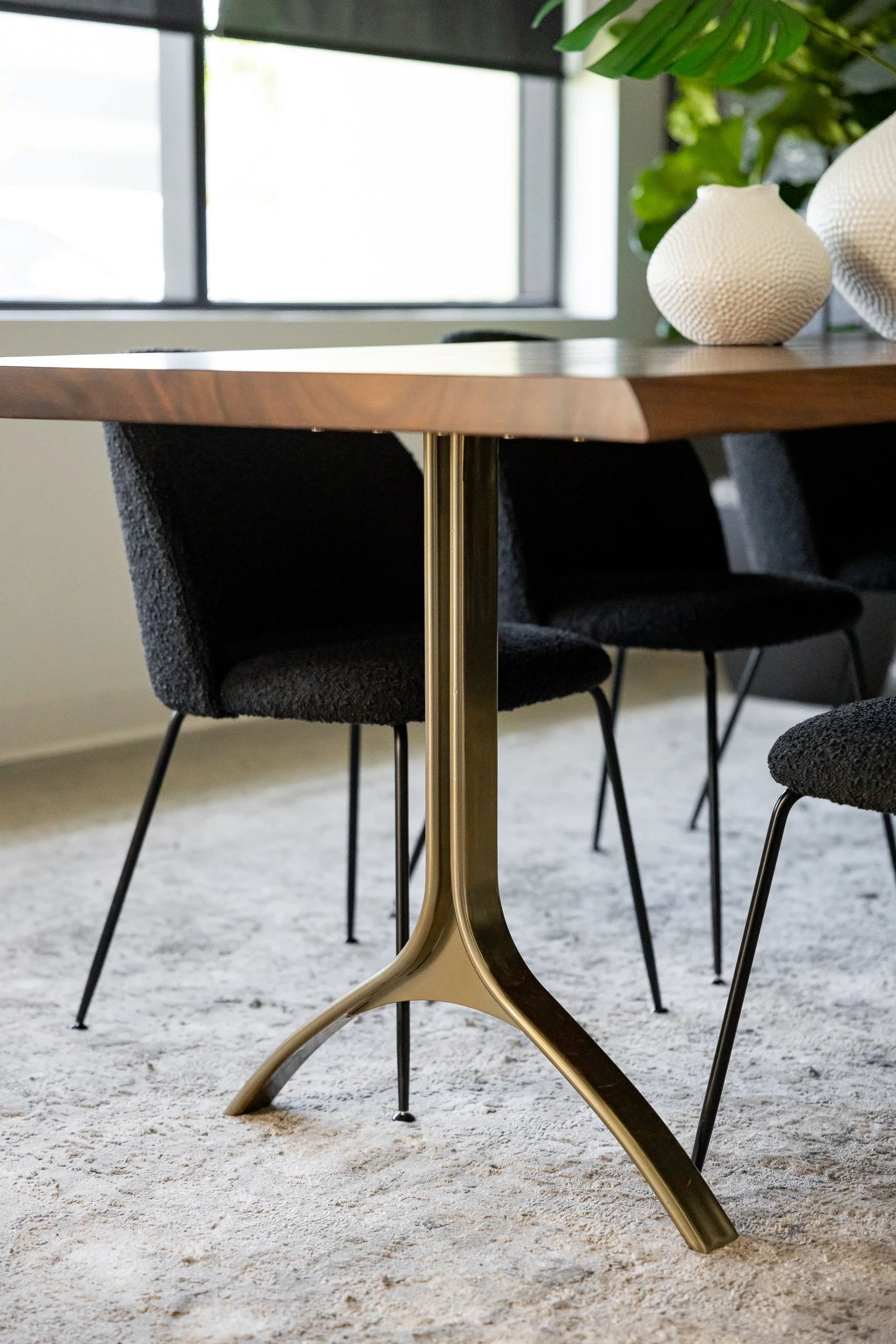 Close-up of a wooden dining table with black chairs. The table has decorative white ceramic vases and a green plant in the background near a window.
