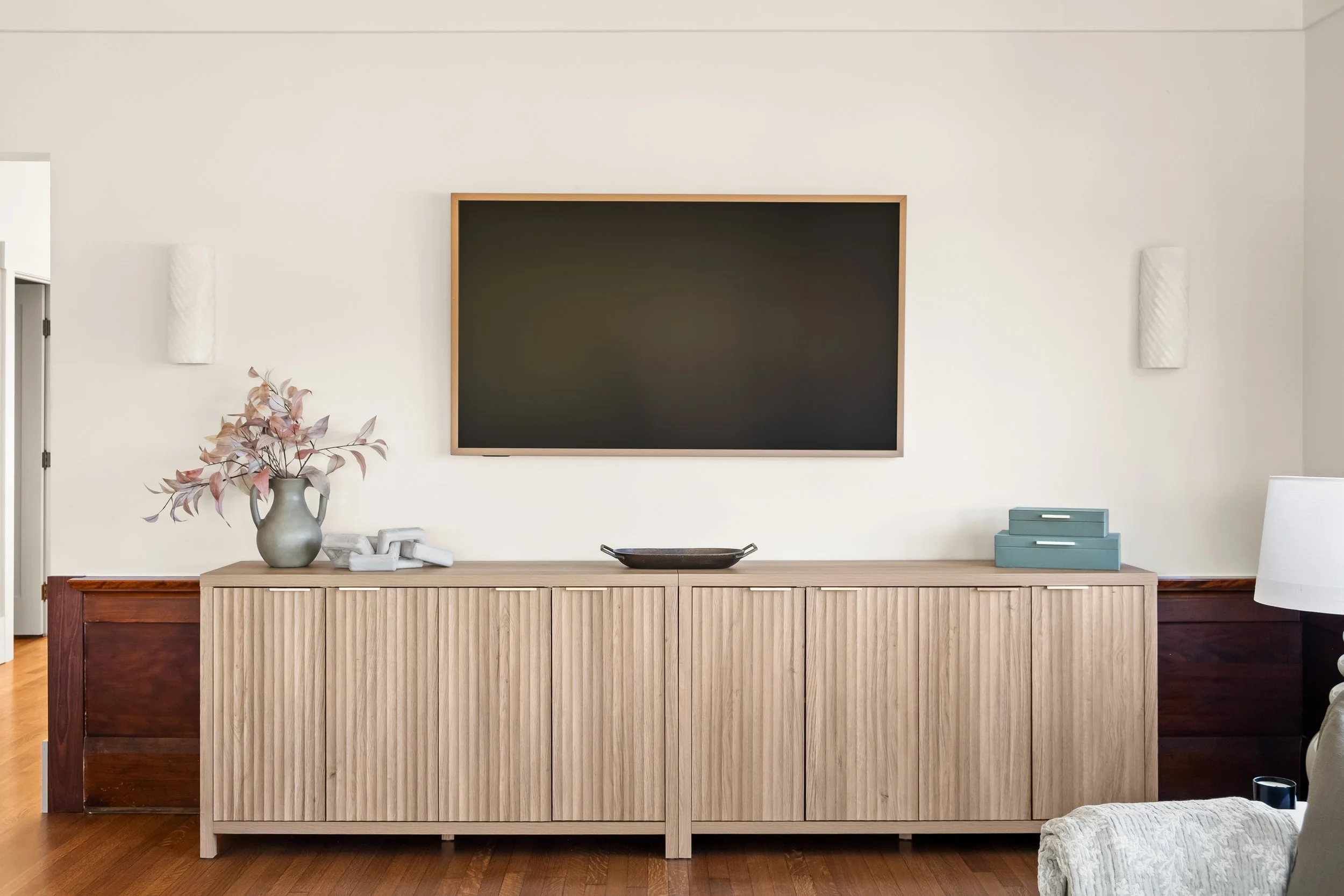 Modern living room with beige and wood-tone furniture, a black-framed chalkboard on the wall, a cream-colored vase with pinkish dried flowers on a sideboard, and a decorative black tray.