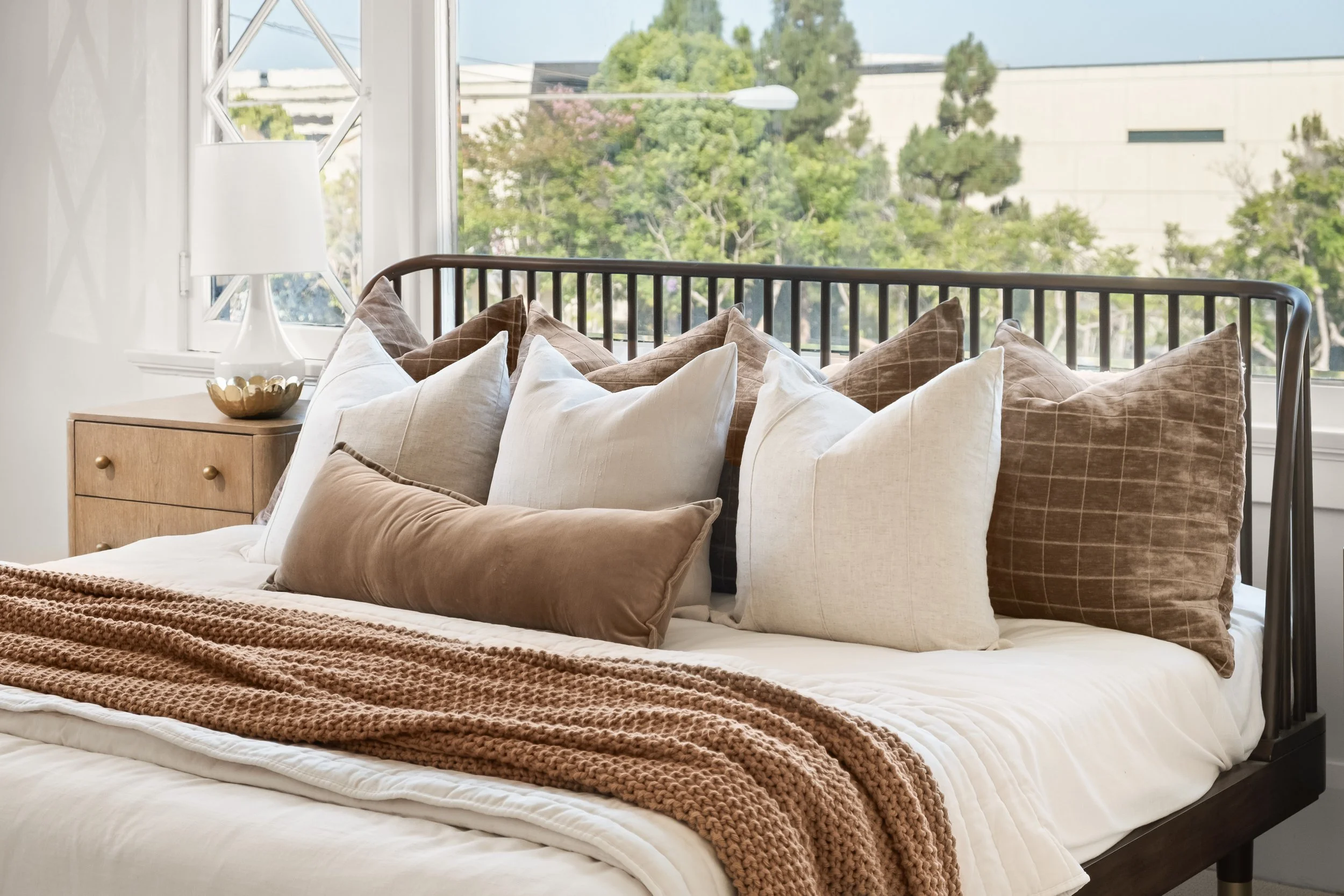 A bed with multiple pillows in various shades of beige and brown, a knitted blanket, a wooden nightstand with a decorative bowl, and a large window showing green trees and buildings outside.