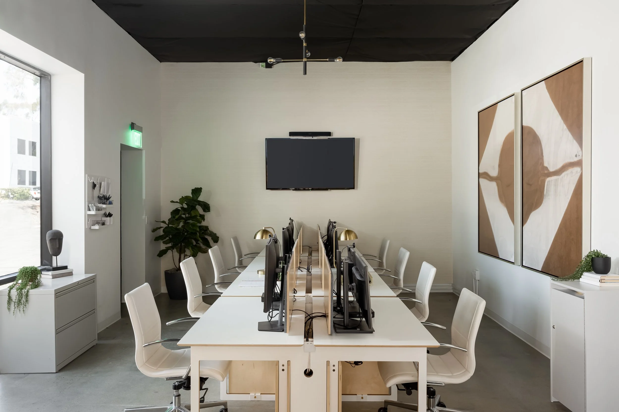 Modern office conference room with white chairs, large windows, a wall-mounted TV, abstract artwork, and a black ceiling.
