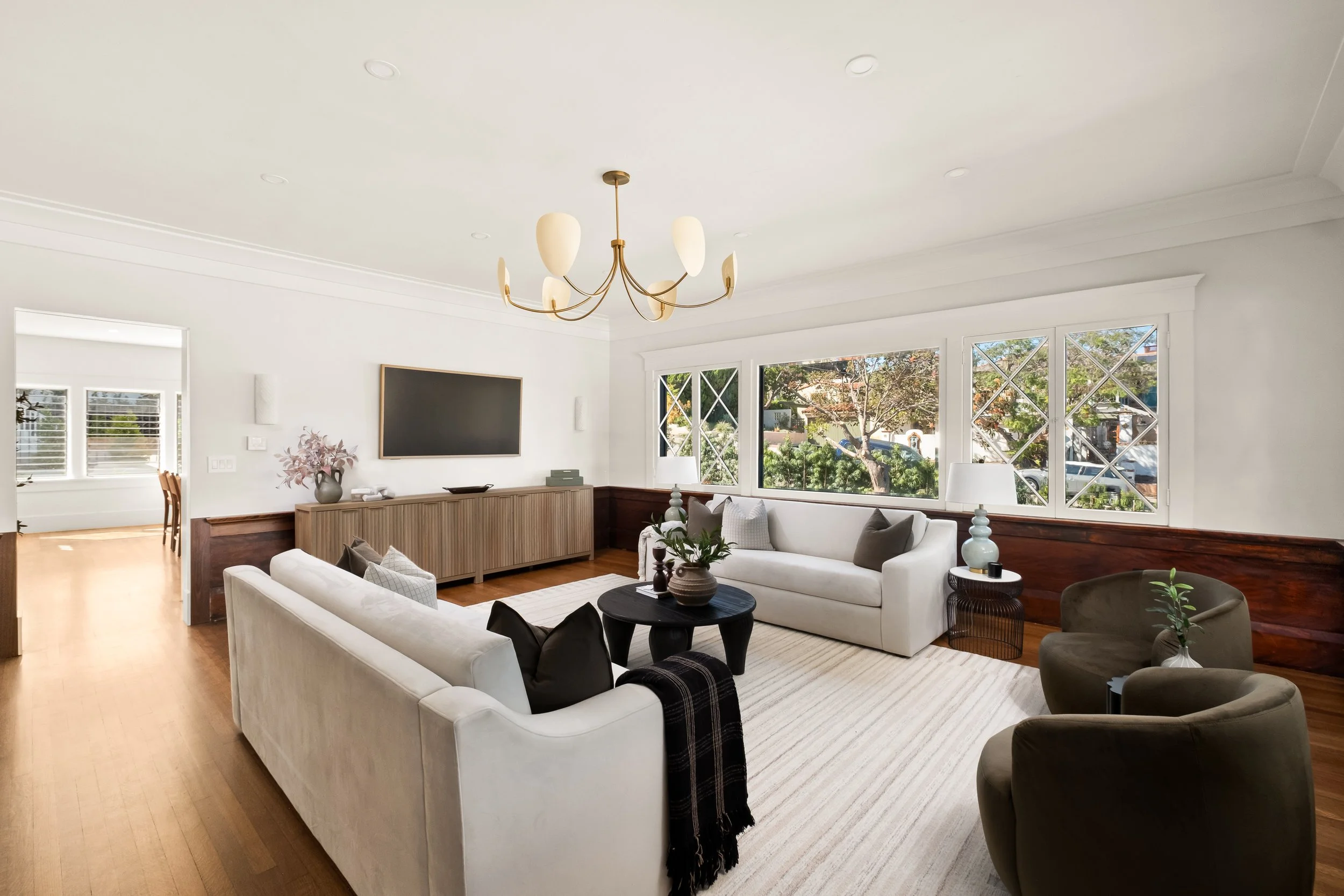 Bright living room with white sofas, a black coffee table, large windows with a view of trees, and a modern chandelier.