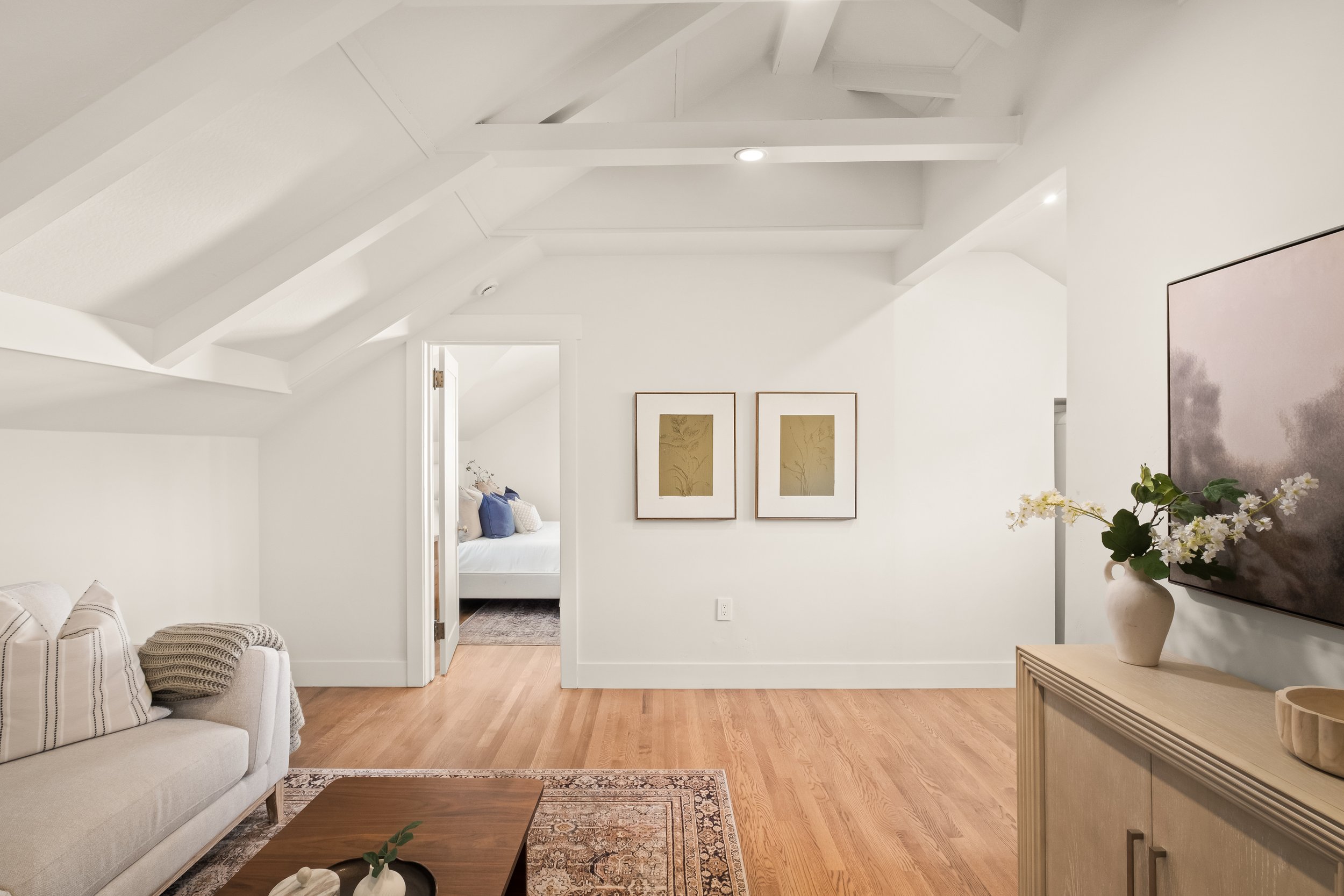 A bright, modern living room with white walls and ceiling, hardwood floors, a beige sofa with pillows, a wooden coffee table, framed artwork on the wall, a dresser with a vase of white flowers, and a large flat-screen TV.