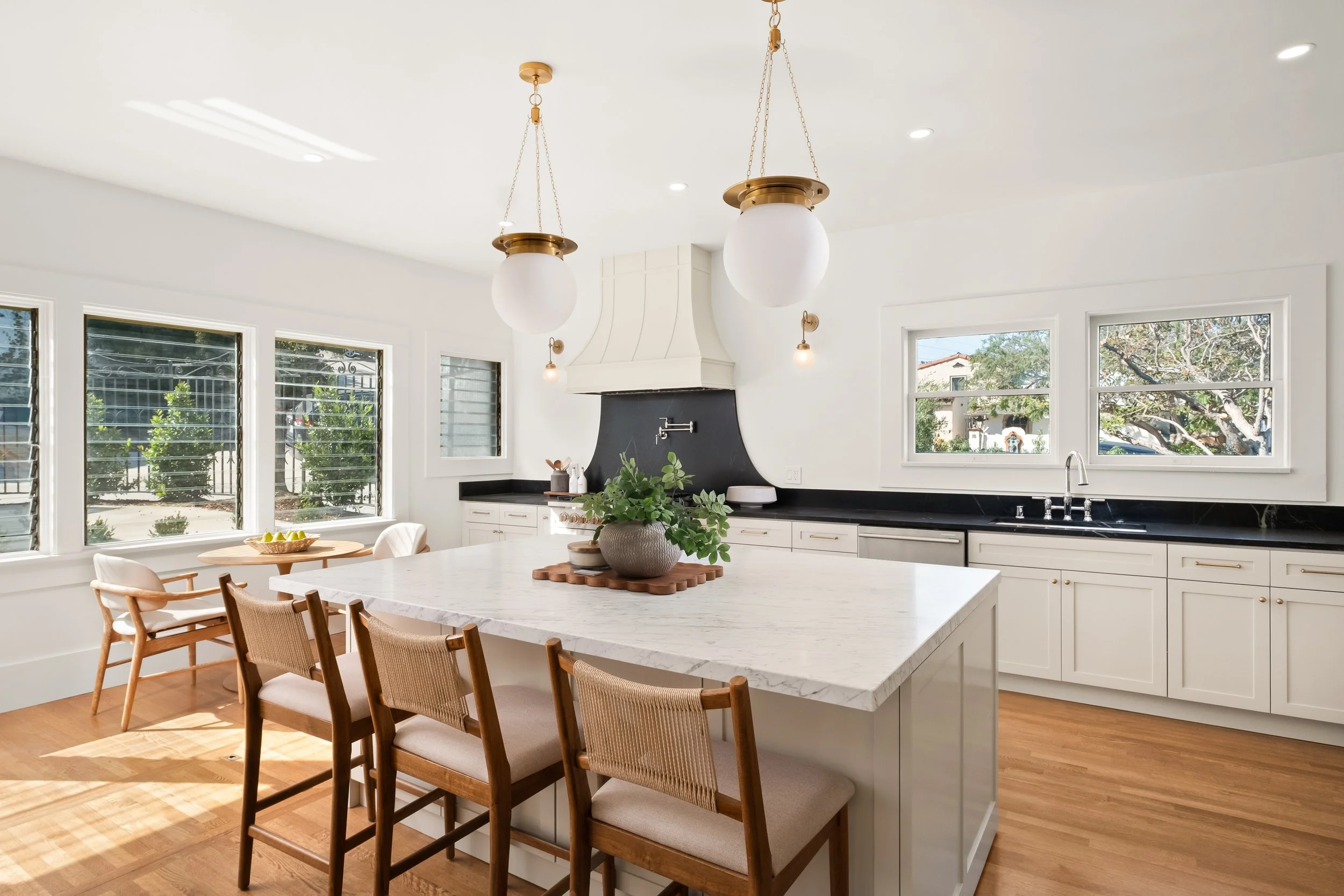A modern kitchen with white cabinetry, black countertop, large windows, and a spacious white marble island with a potted plant. Two gold pendant lights hang from the ceiling.