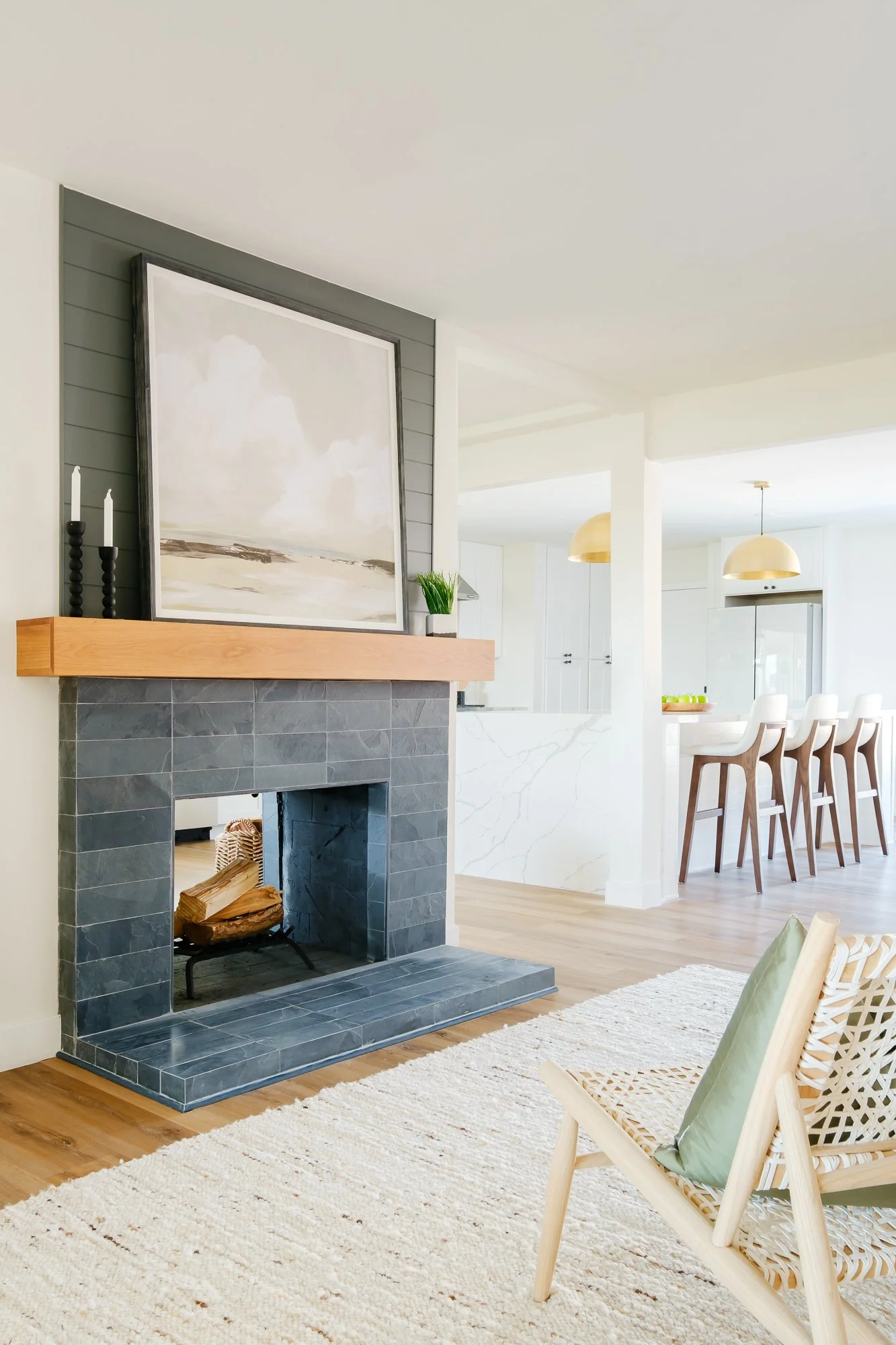 A modern living room featuring a fireplace with a wooden mantel and charcoal tiles, a large abstract painting, a cream-colored rug, light wood flooring, a woven armchair with a green cushion, and a bright, open kitchen with white cabinetry, bar stool