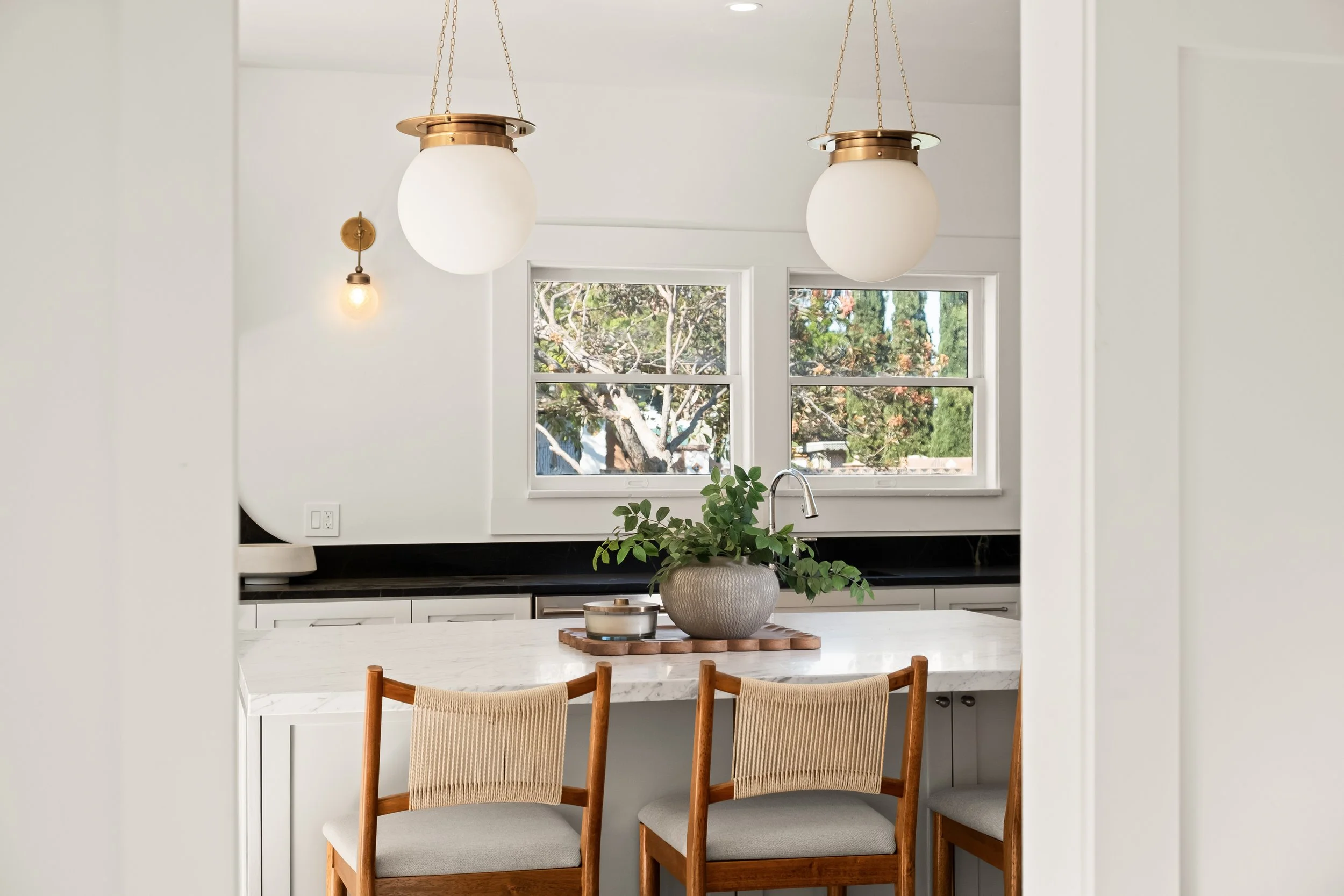 Kitchen view with two pendant lights, two windows showing trees outside, a pot with a green plant on the countertop, and wooden chairs at a white marble table.