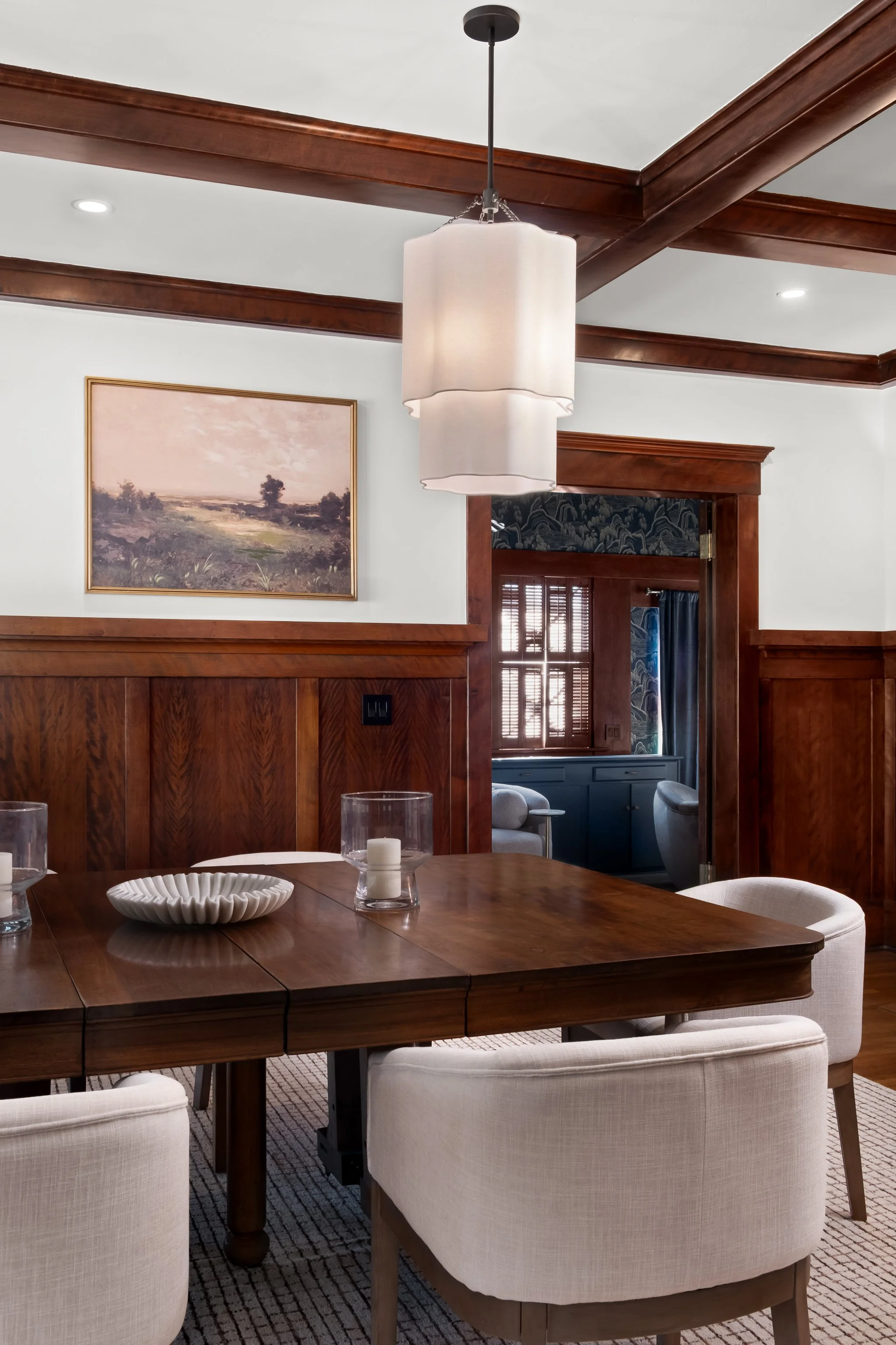A wooden dining table with white upholstered chairs, decorative candles, a white bowl, and glassware, in a room with wood trim, a painting, and a doorway leading to a sitting area.