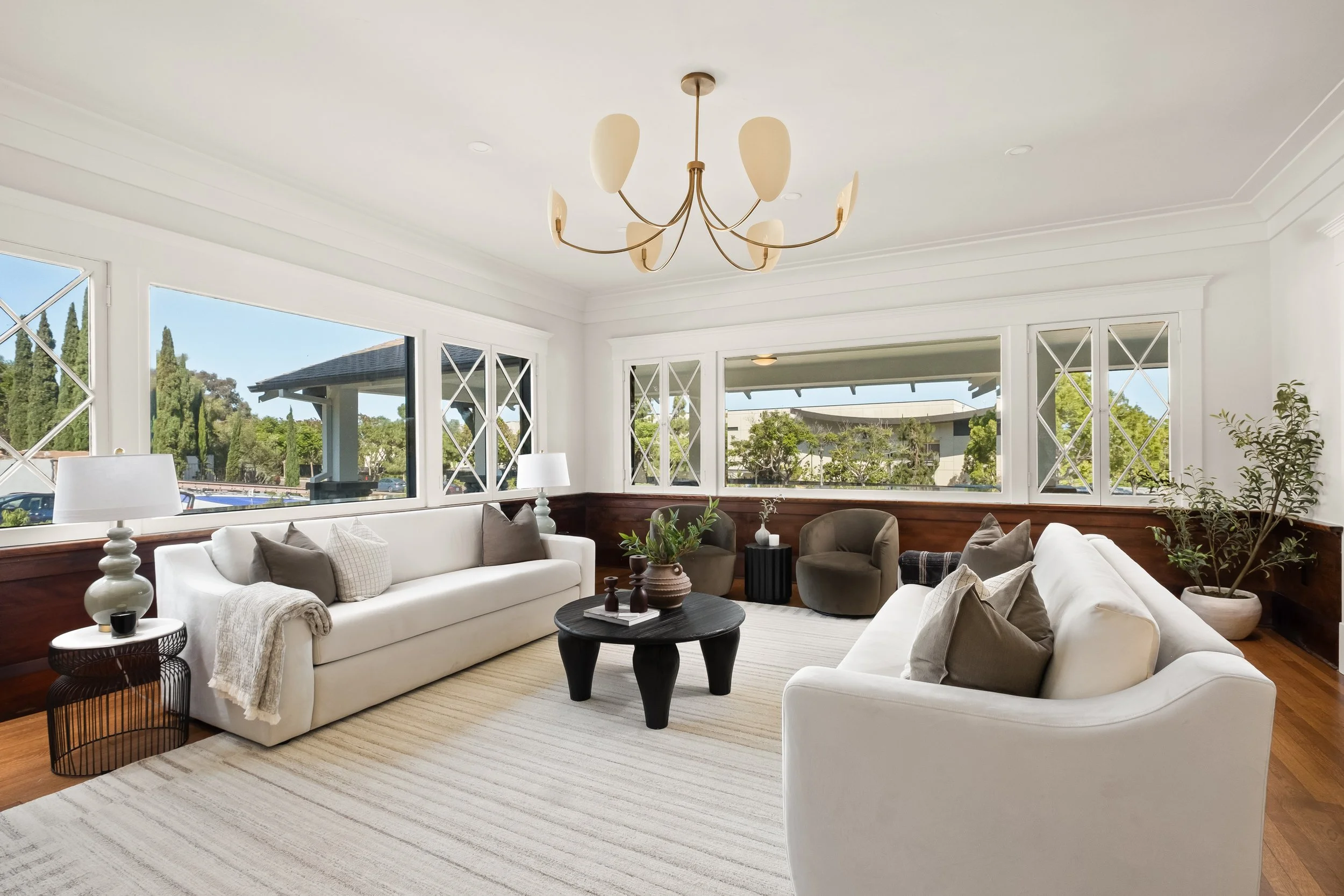 Elegant living room with white sofas, green armchairs, a black coffee table, modern lamps, and large windows with a view of greenery outside.
