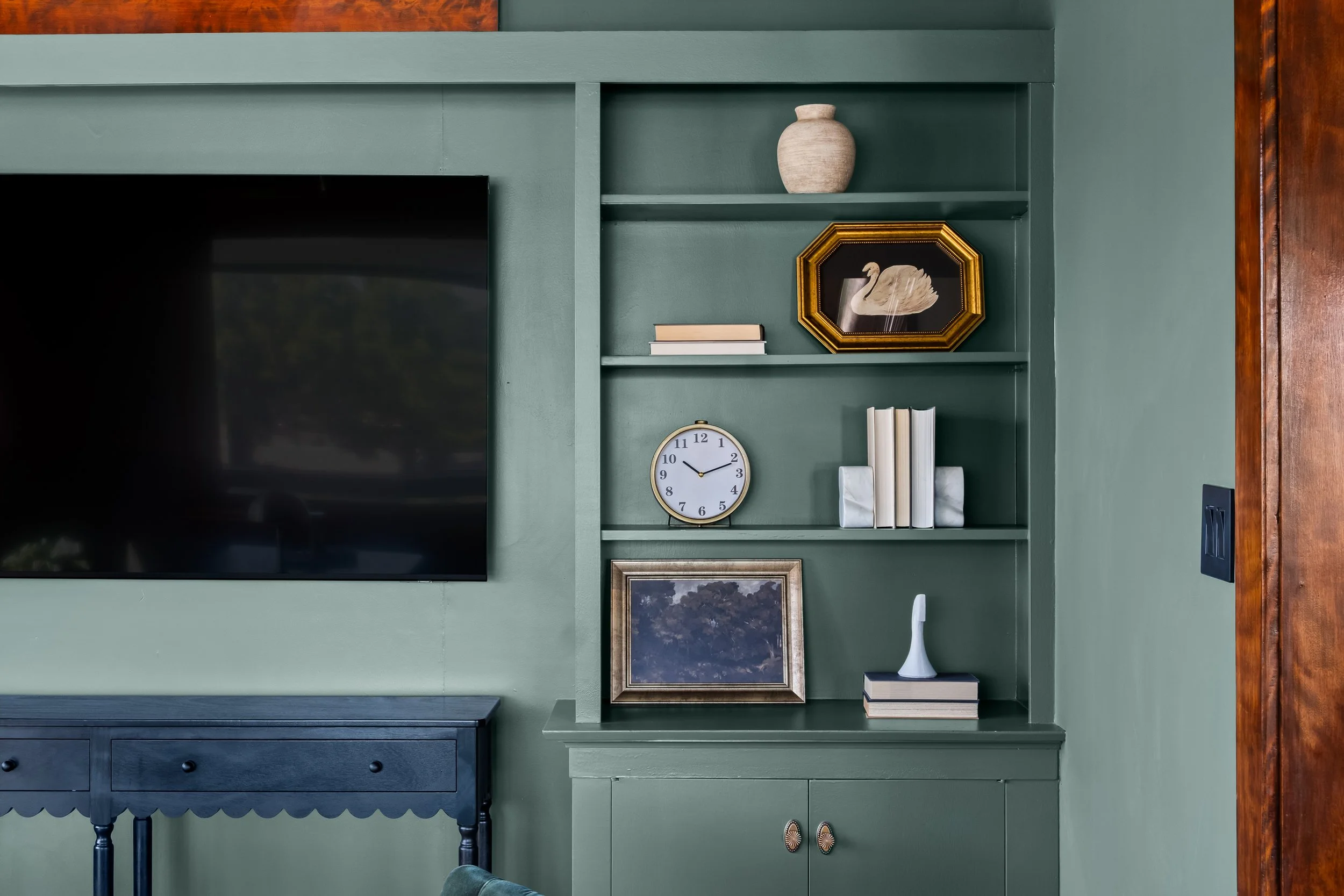 Green built-in shelving unit with decorative items including a beige vase, a framed swan picture, a clock, and books. A large flat-screen TV is mounted to the left. There is a dark blue table below the TV and part of a green upholstered chair in the 