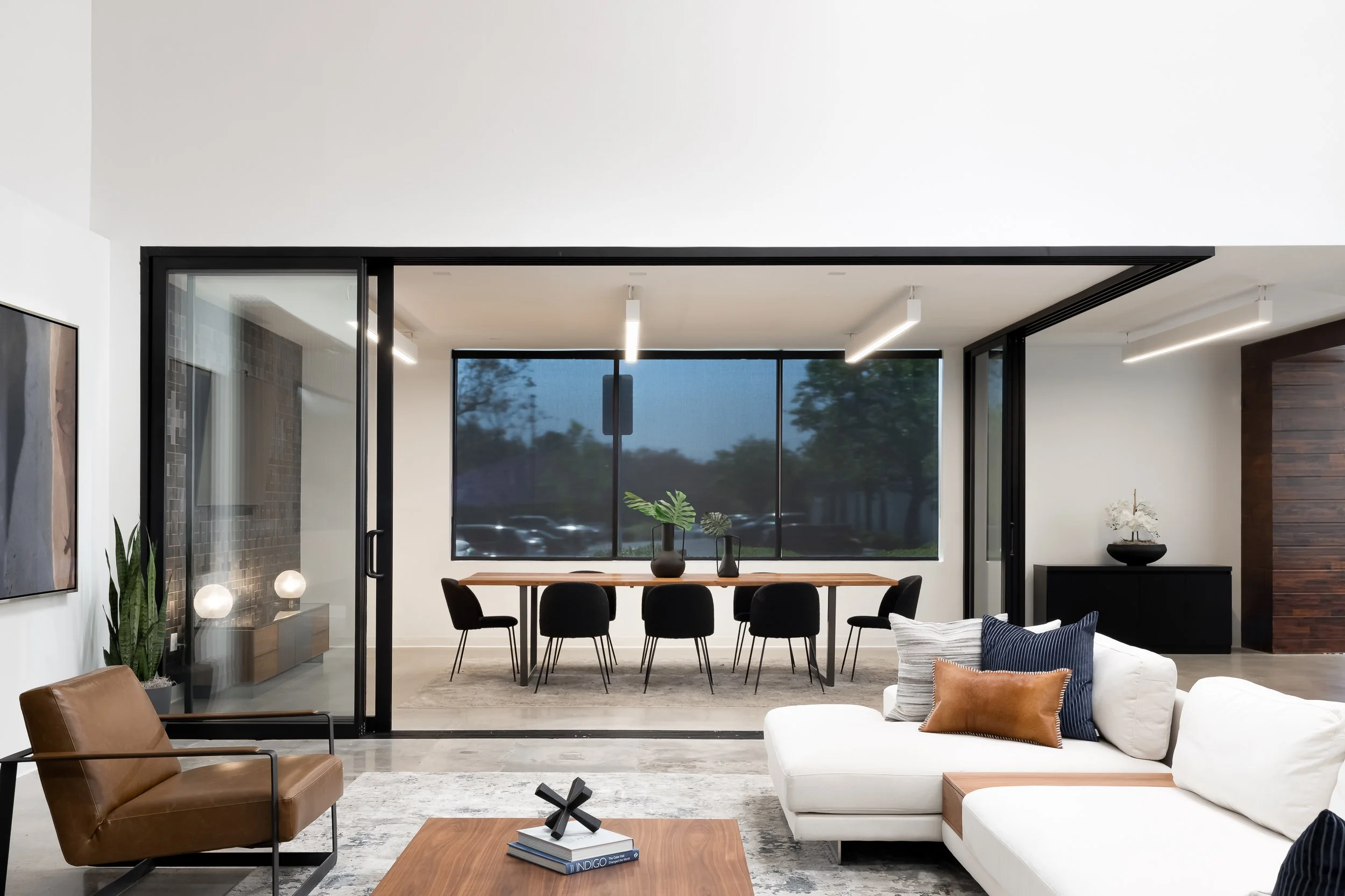 Modern living room with white walls, black framed glass doors, a wooden dining table with black chairs, and a seating area with white sofa, brown armchair, and decorative pillows, with large windows showing trees outside.