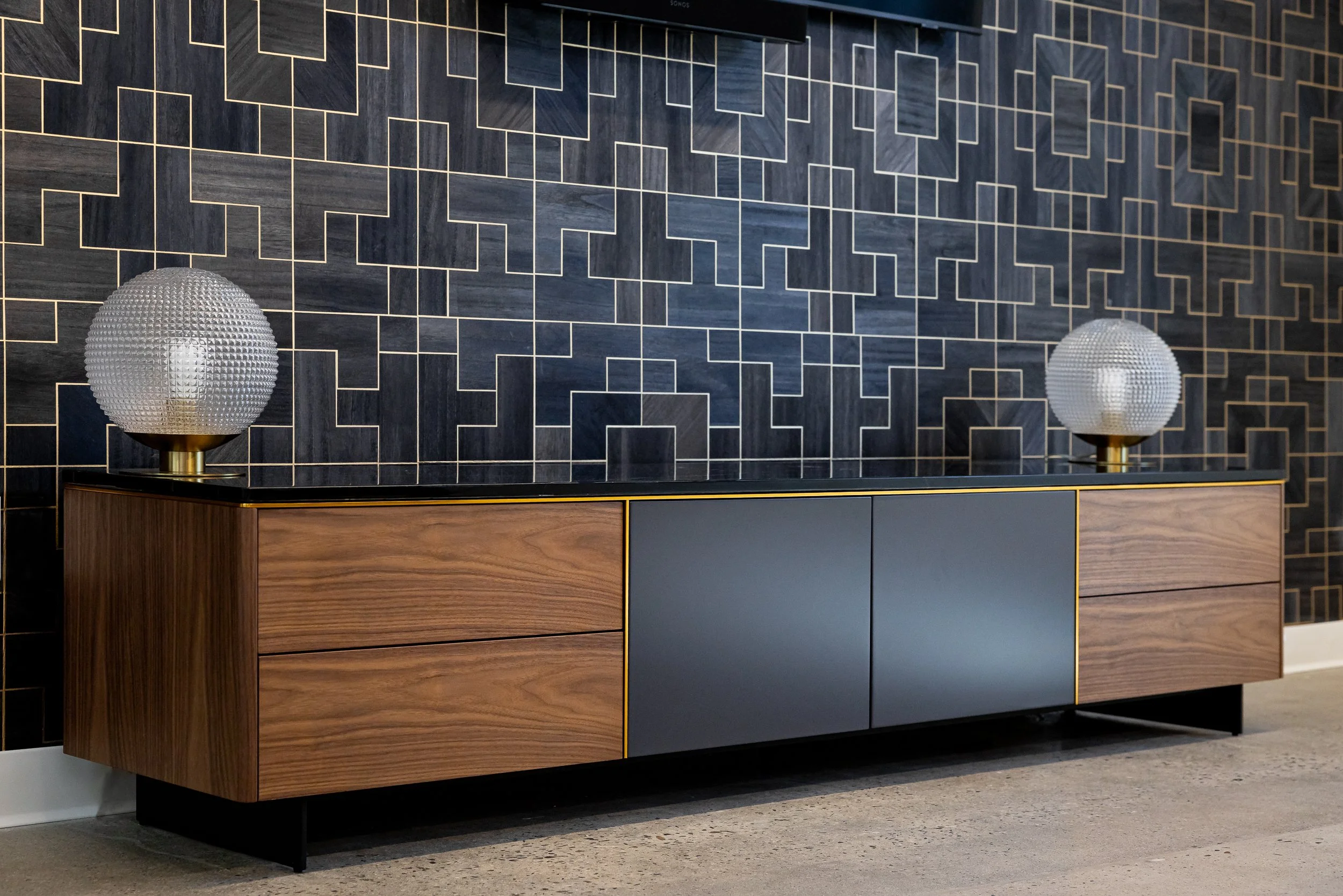 A modern sideboard with wooden and dark gray panels, topped by two white textured table lamps with gold bases, set against a dark wall with geometric gold line patterns.