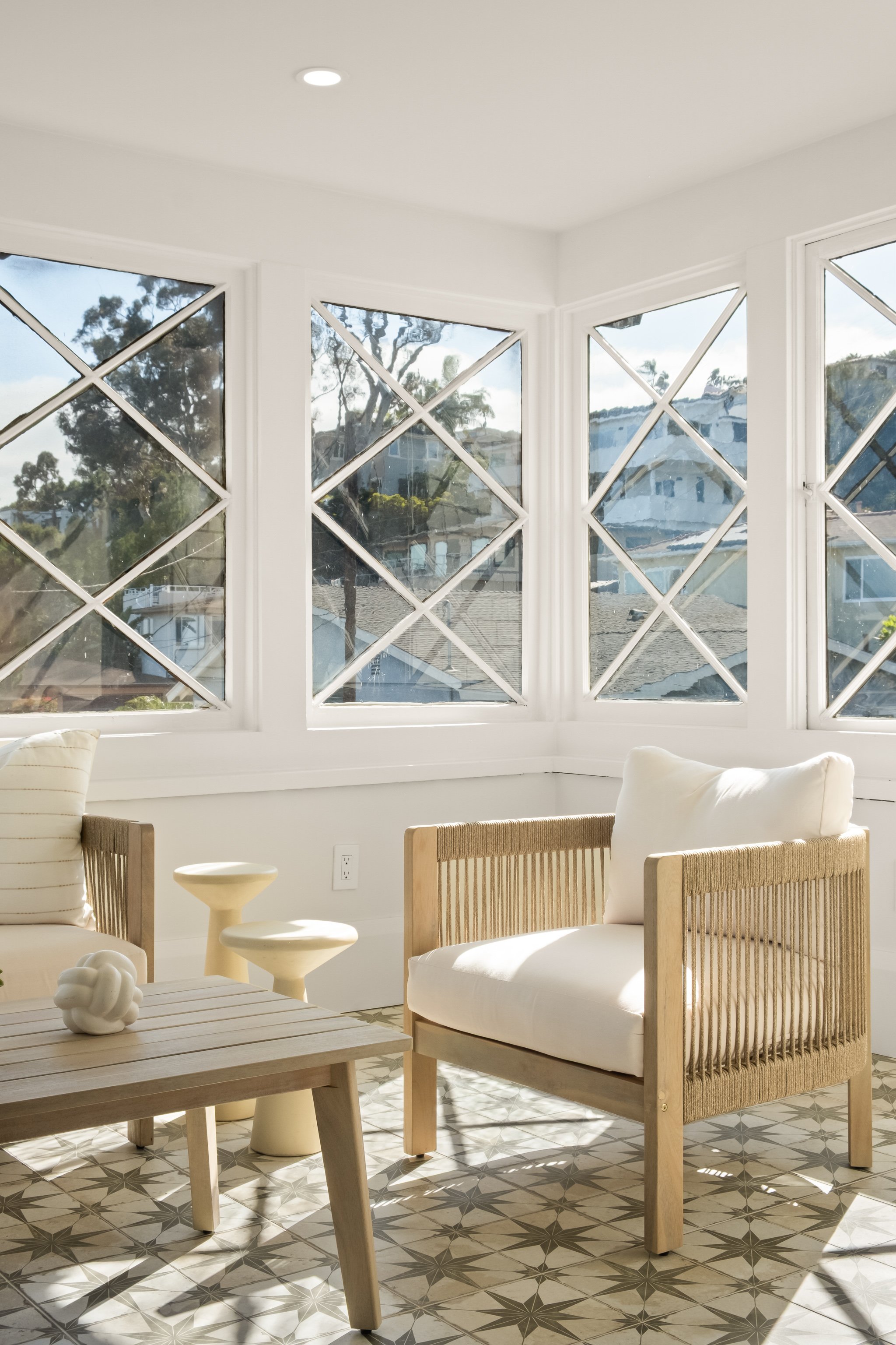 A bright living room with a large bay window featuring diamond-shaped panes, white walls, patterned floor tiles, wooden furniture, and neutral-colored cushions.