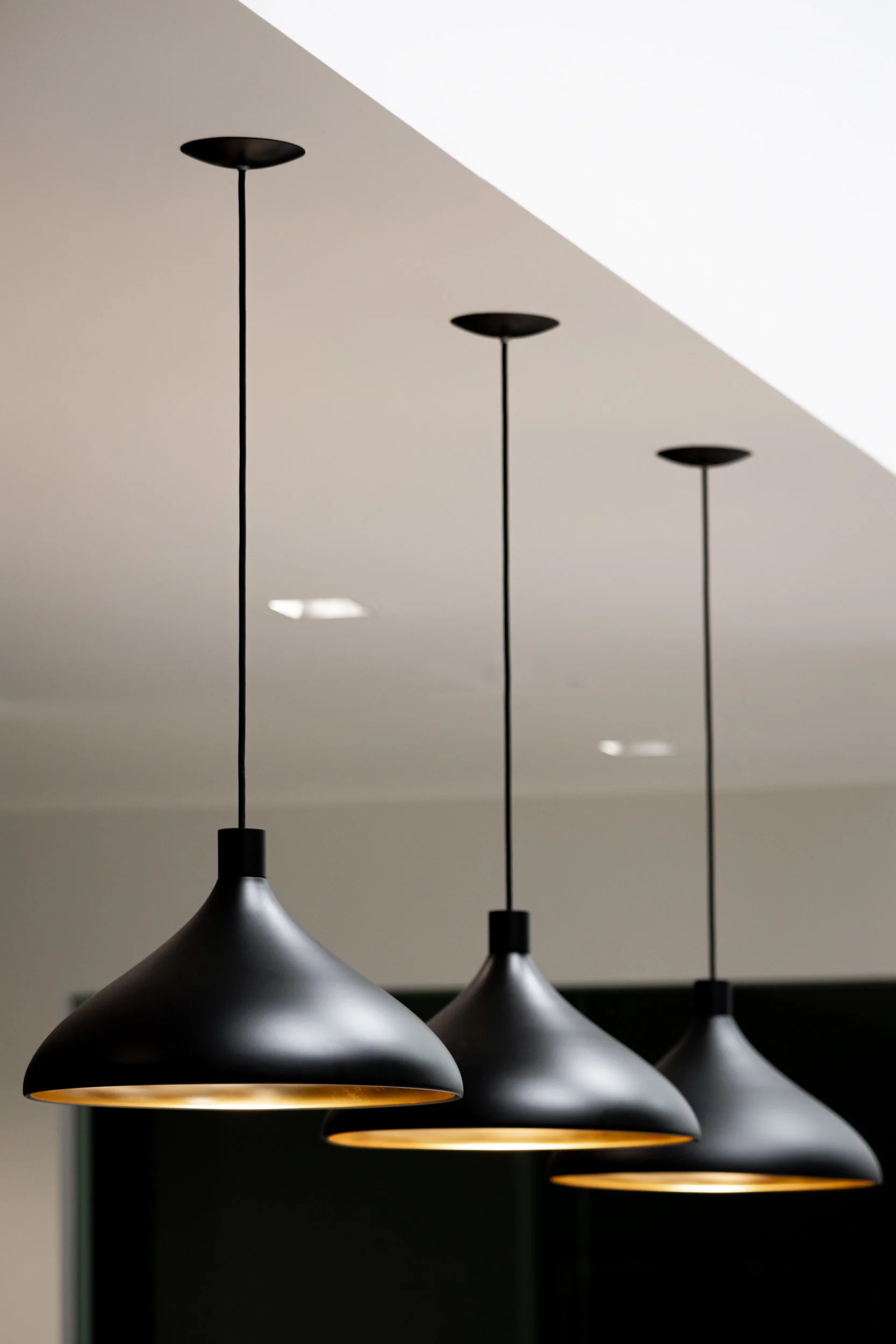 Close-up of several modern black pendant lights hanging from the ceiling with a white background and a dark wall below.