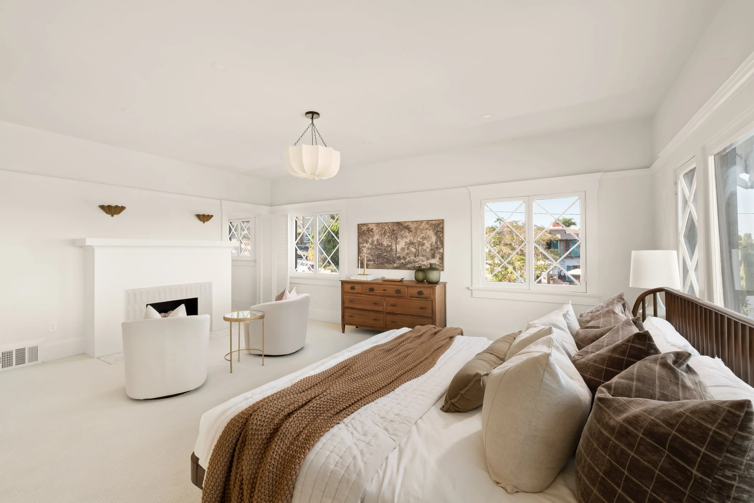 A bright, white bedroom with large windows, a wooden dresser, a bed with white bedding and brown accents, and modern decor.
