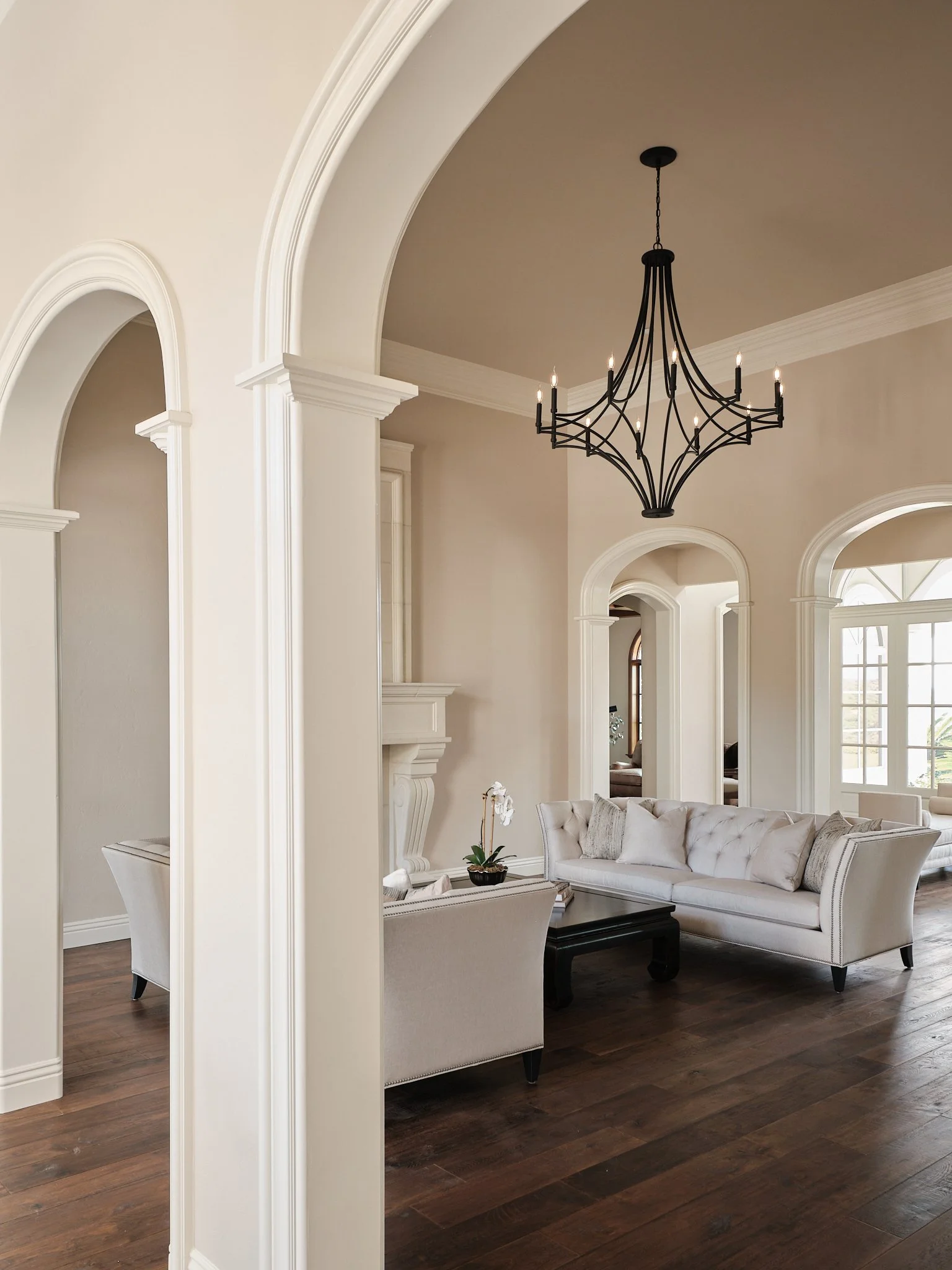 An elegant living room with high ceilings, white walls, large windows, and a black chandelier. There is a white sofa with decorative pillows and a small table with a plant.