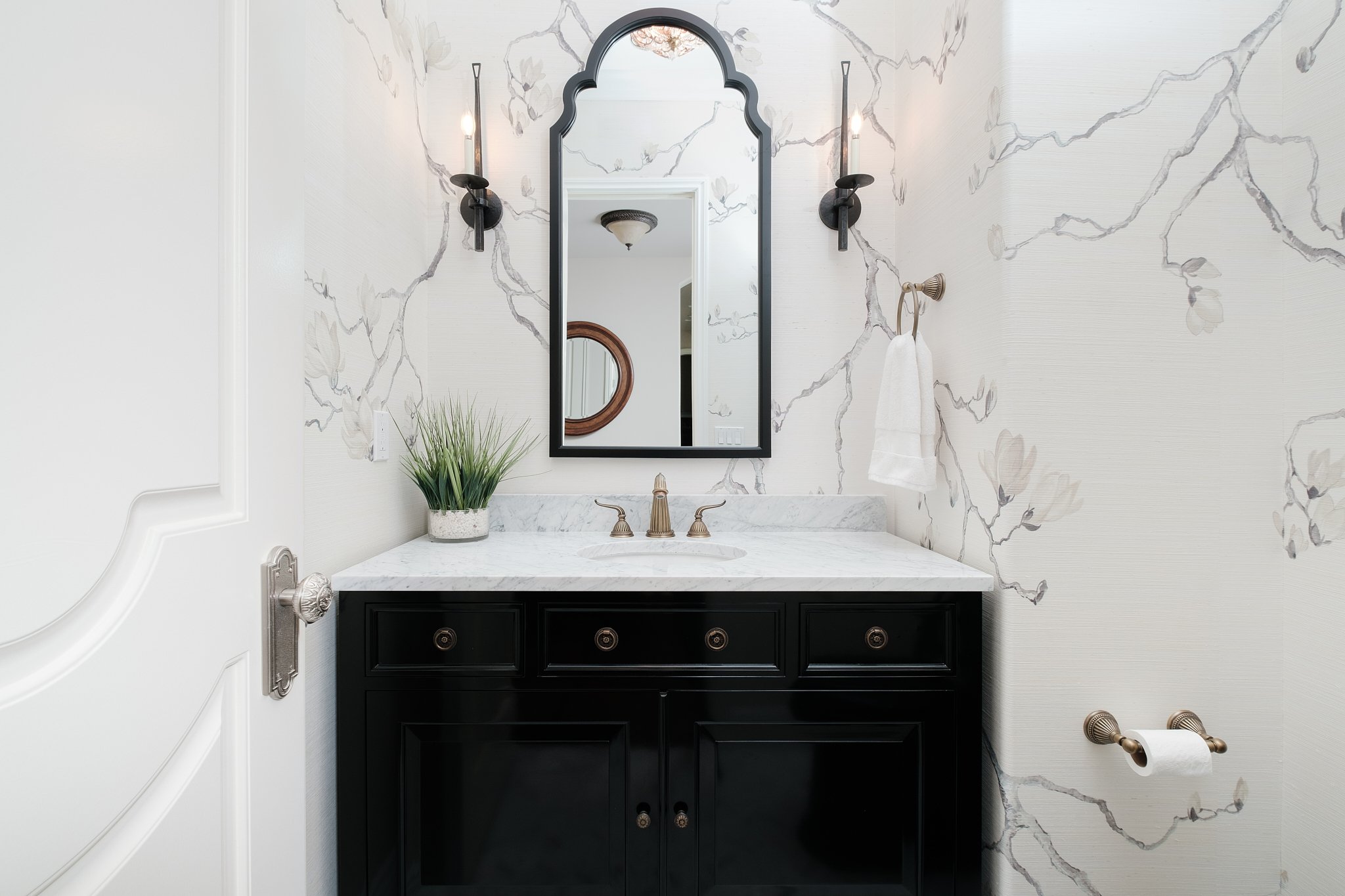 Bathroom vanity with a black cabinet, white marble countertop, and a modern faucet. A large, arched black-framed mirror hangs on the wall, which has a wallpaper with a floral and vine design in soft gray tones. A small green plant is placed on the co